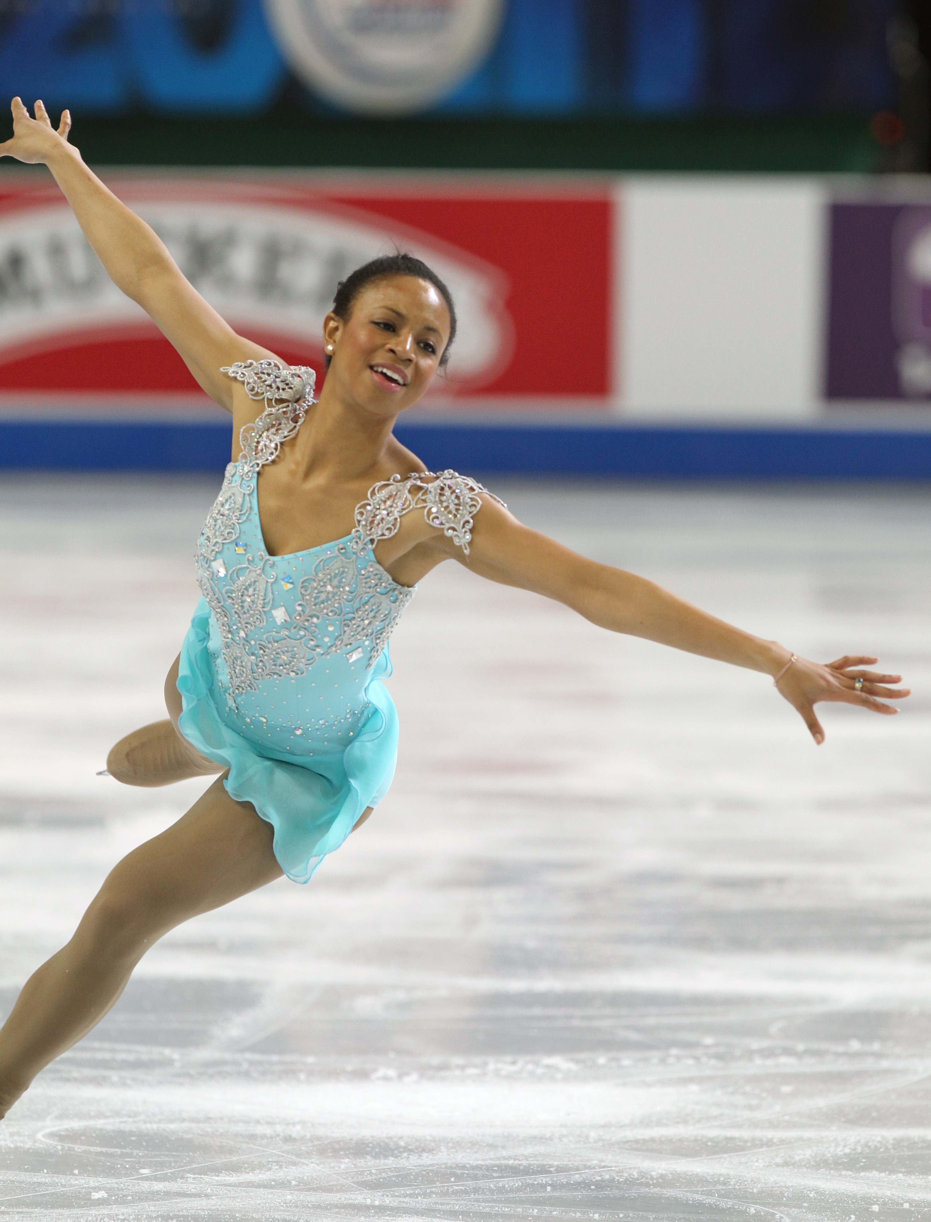 Musademba skates wearing a light blue skating costume with silver accents on the shoulder. She stands with both arms extended outward and one leg extended behind her