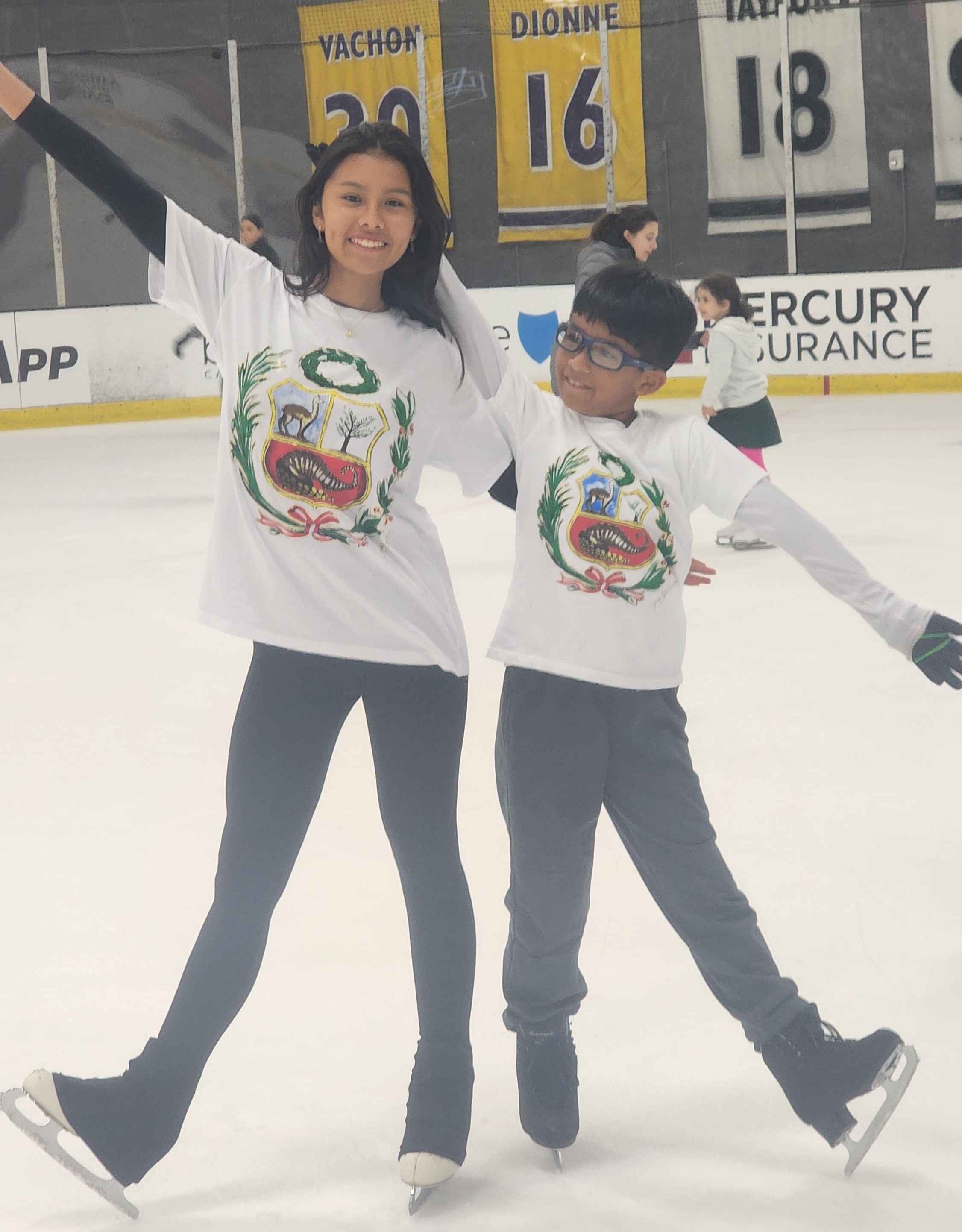 Valerie and Sebastian Aguilar pose on the ice with one arm around each other and the other extended outward. Valerie (left) is a young Hispanic woman with long black hair, and Sebastian is a young Hispanic boy with shirt black hair and glasses. They are both wearing the same tshirt with a multi0colored shield design. 