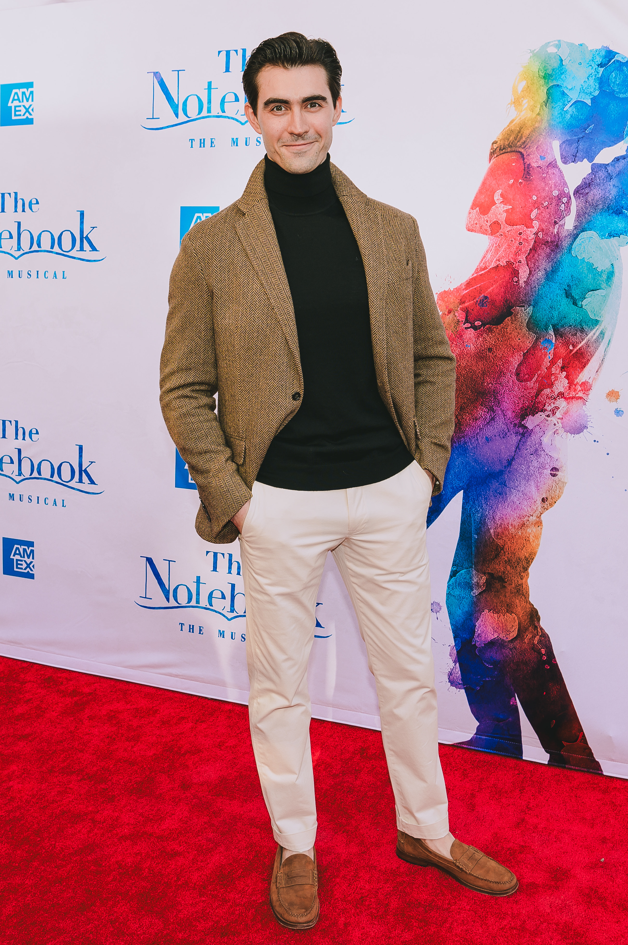 Alex Benoit, in a black shirt, brown jacket and beige pants, stands on the red carpet for the Notebook.