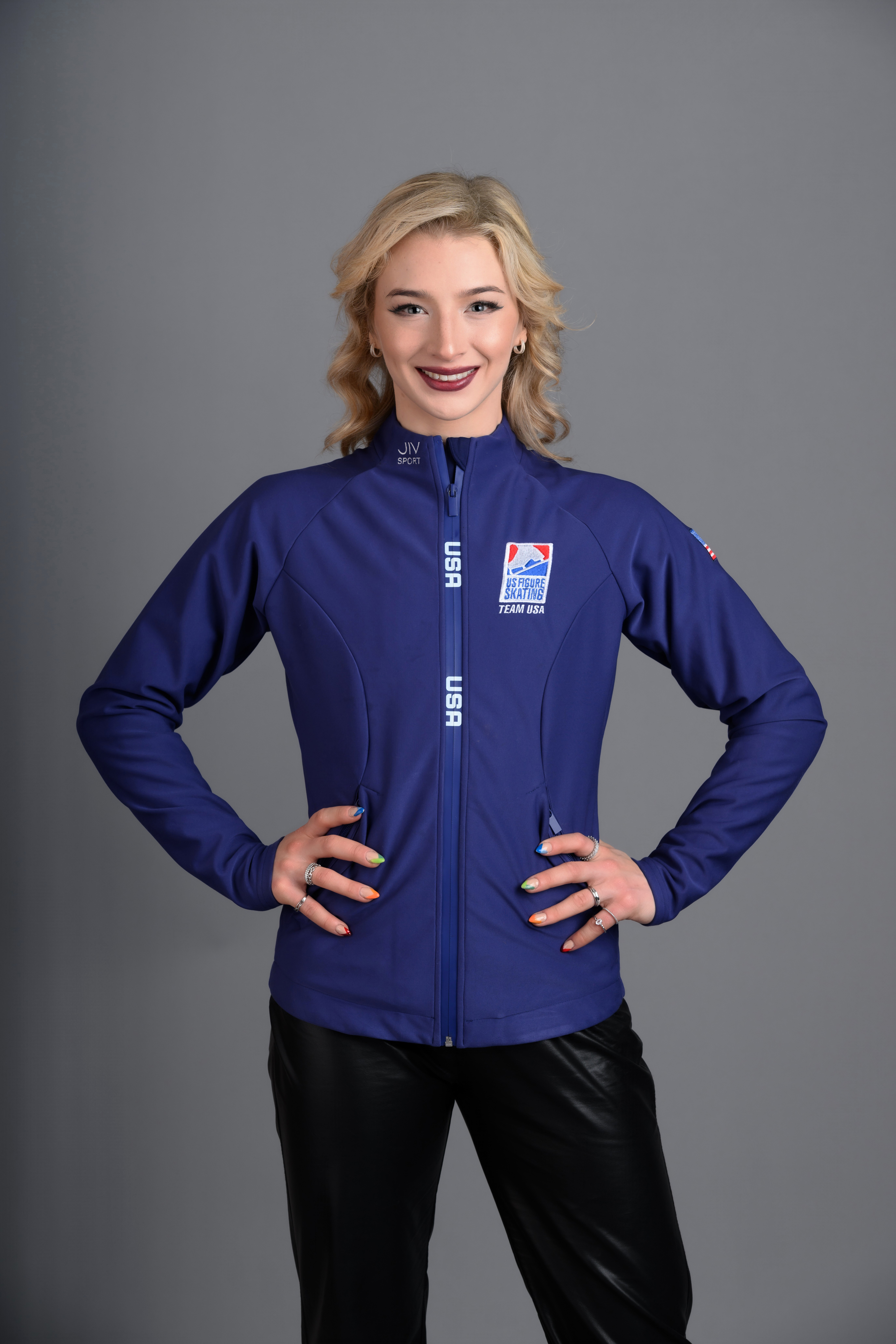 Amber Glenn, with a smile, wearing her Team USA jacket, with hands on hips.