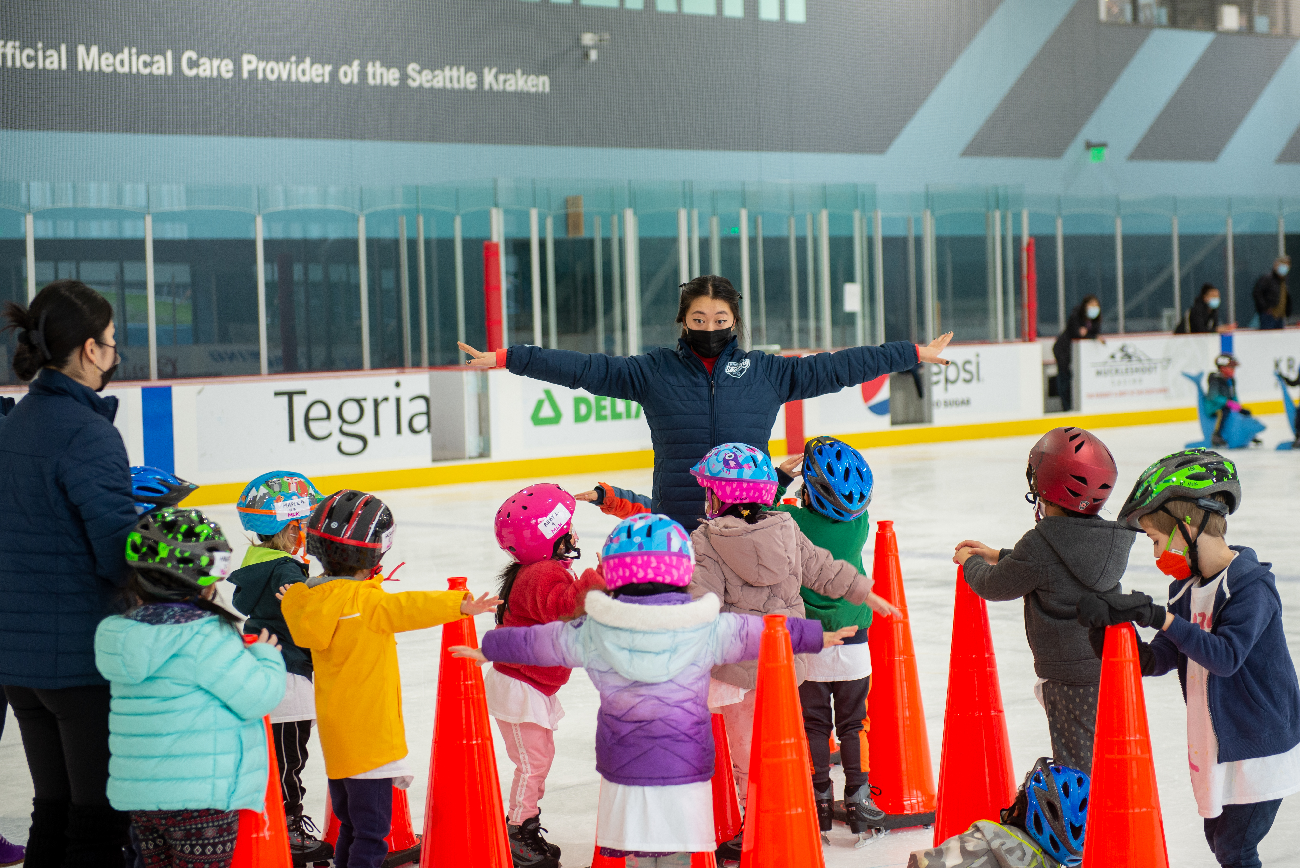 A group of young learn to skate skaters stand in front of an instructor with their arms out wide.