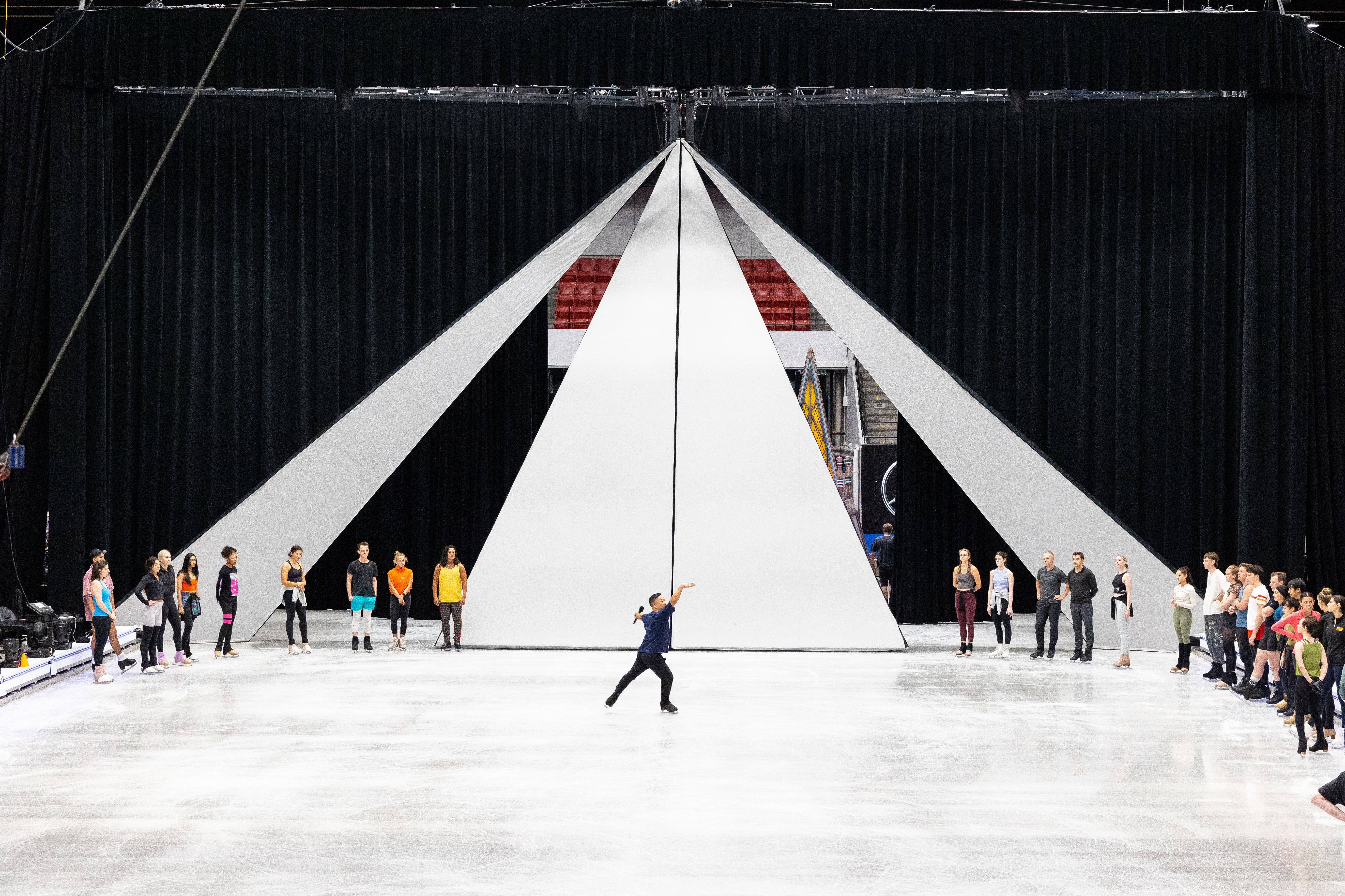 Gig Siruno skates with a mic in his hand, the Disney on Ice cast standing in a circle around him watching him.