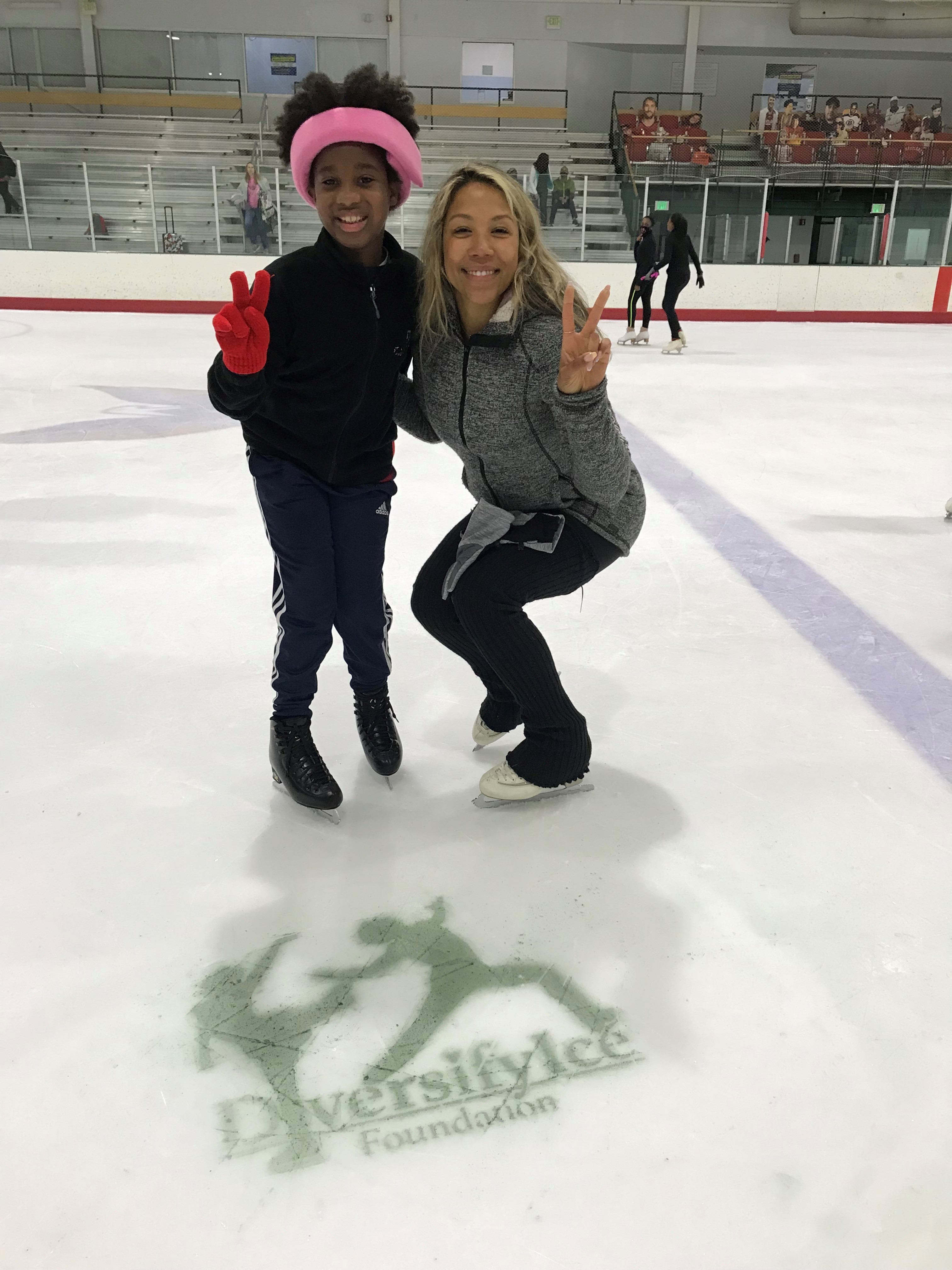 Photo of Megan Williams-Stewart posing with a young female skater. She is wearing a grey jacket and black pants, and the young skater is wearing a black jacket, blue swetpants and a bright pink headband. 