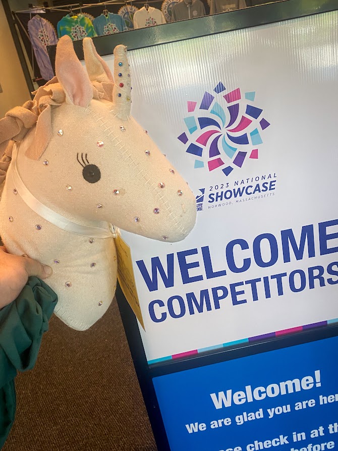 Photo of the Terryl Tossie - a unicorn head plush - in front of a sign that says "Welcome Skaters" and has the logo for the 2023 National Showcase