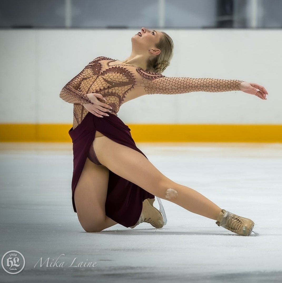 Brooke Tufts kneels on the ice with her left arm extended outward during her performance. She is a woman with blonde hair wearing a brown skating costume