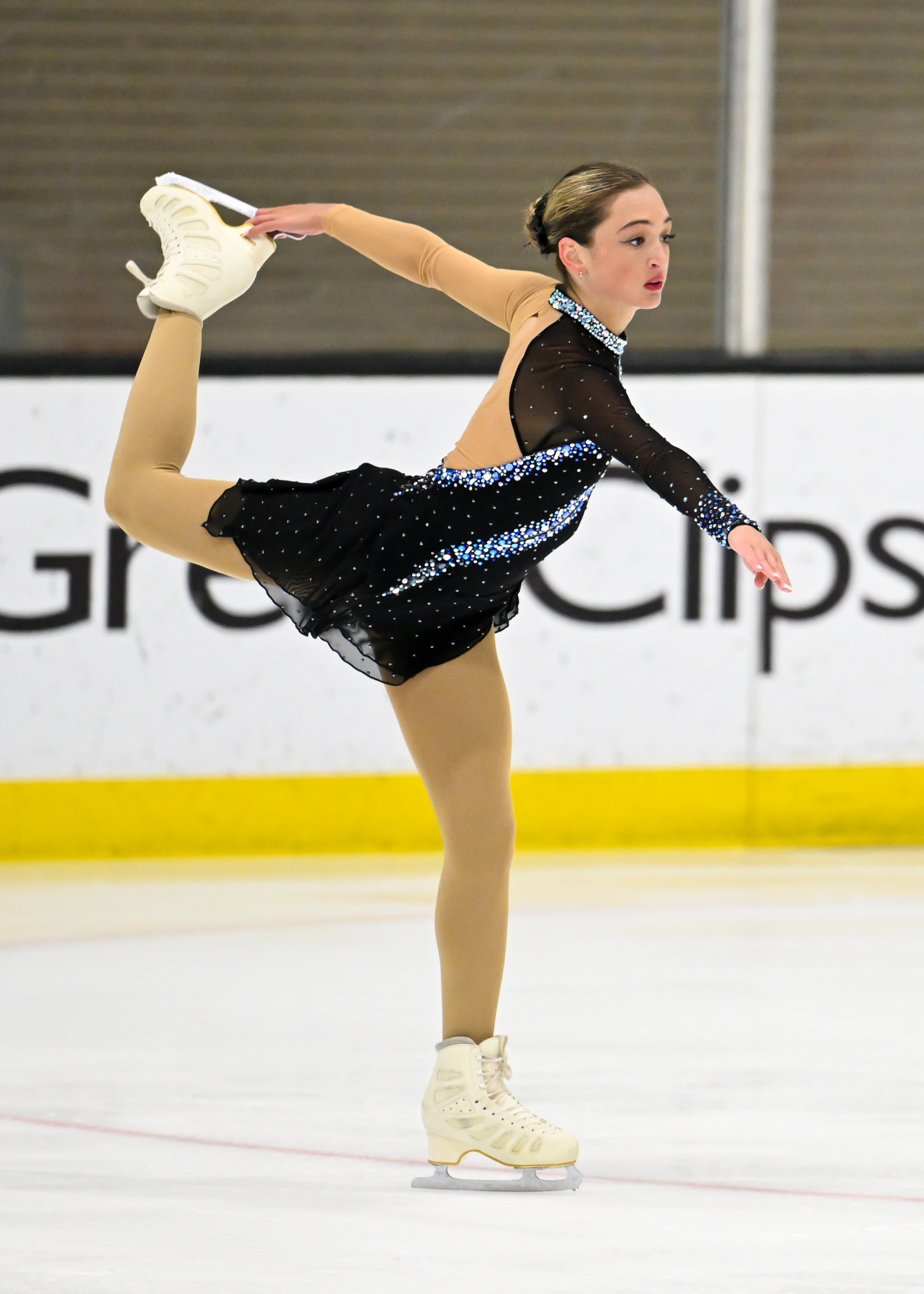 Lindsey Byer skates holding up one leg with her left arm. She is wearing a longsleeve black skating costume with blue gems. He hair is tied up in a bun