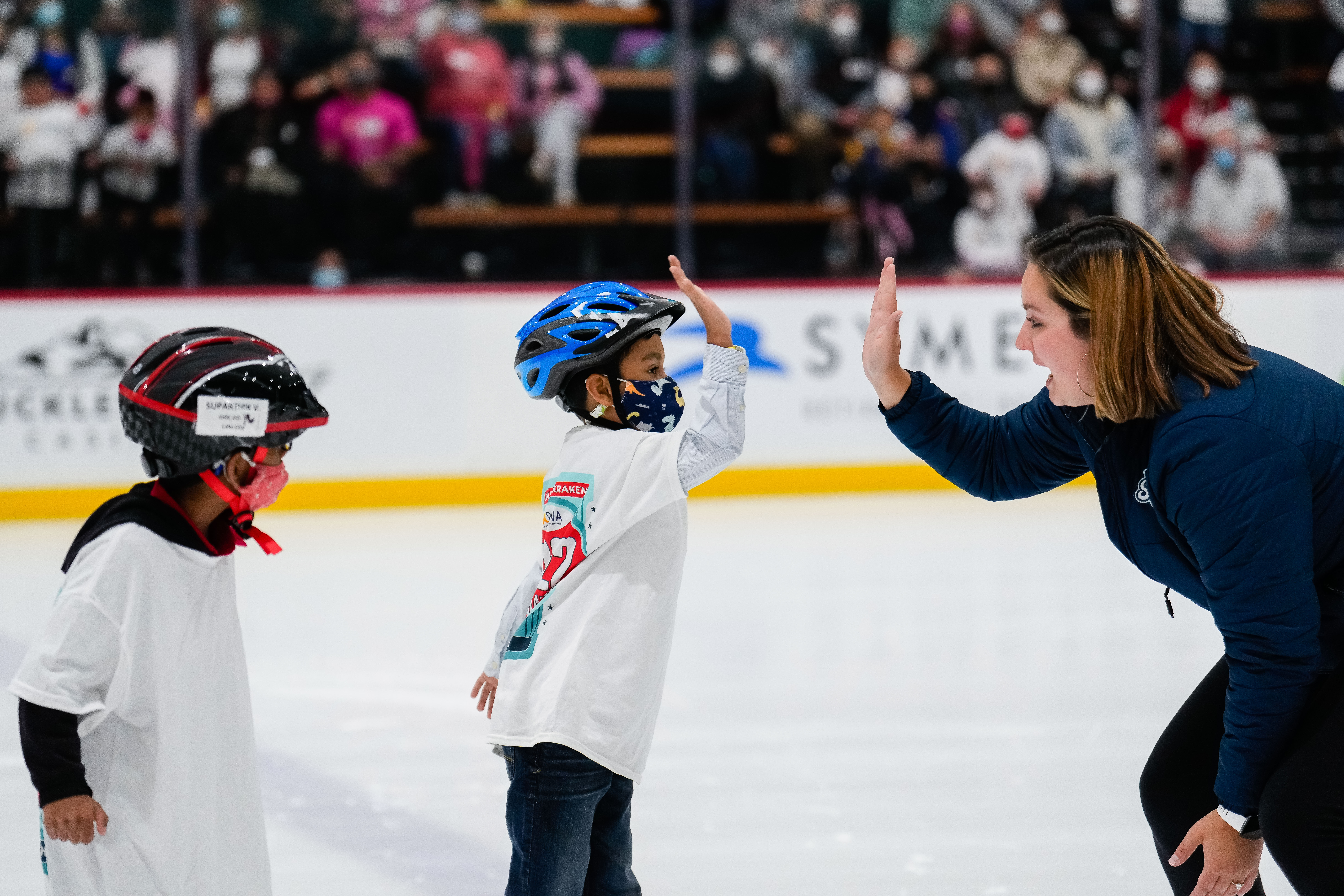 Photo of an instructor and skater sharing a high five. The skater is wearing a large white tshirt over a white long sleeve shirt and a blue helmet. The instructor is bending down and has shoulder length brown hair and a navy blue jacket on with the Kraken logo. 