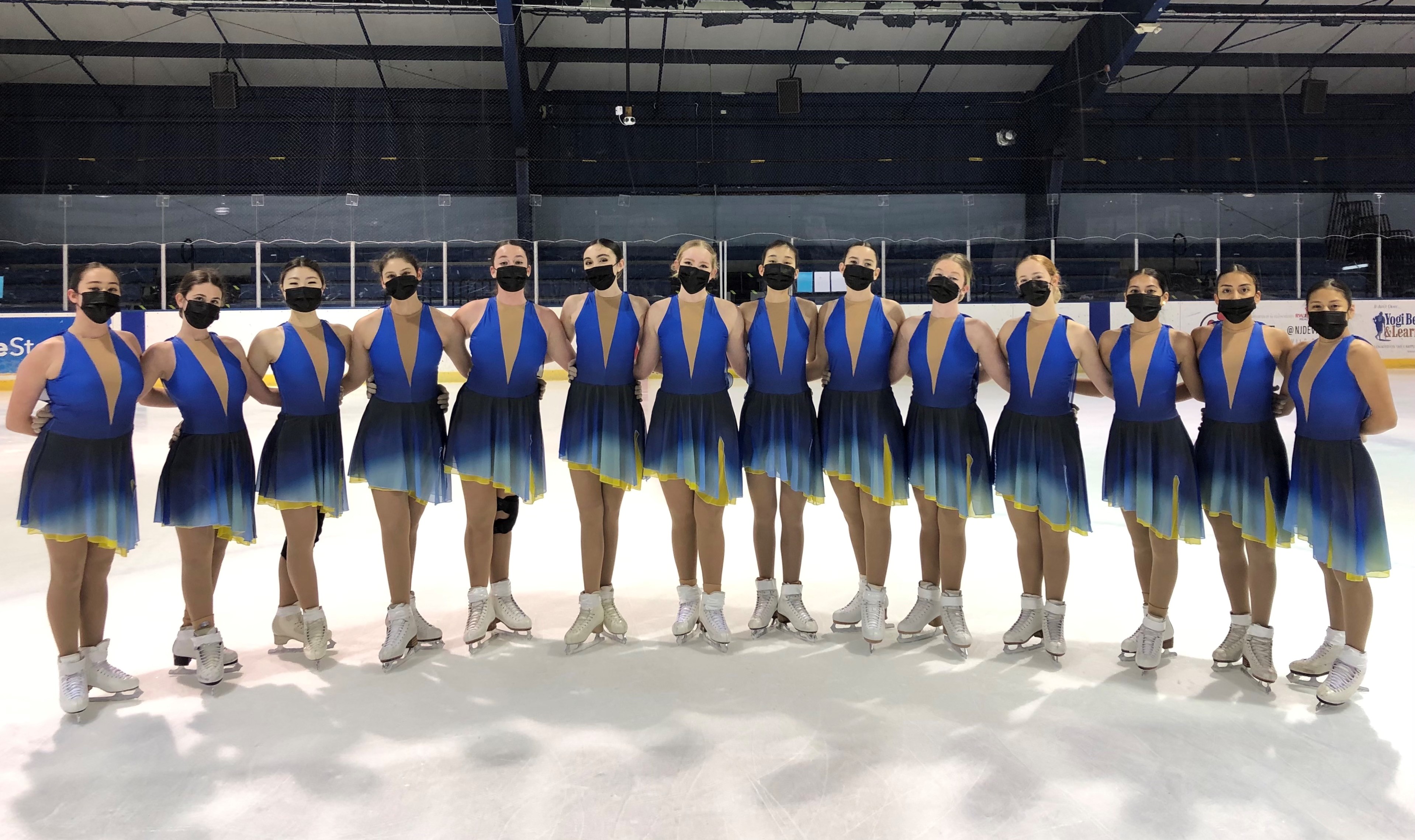 CP Ice skaters line up in blue dresses and black masks for a posed photo.