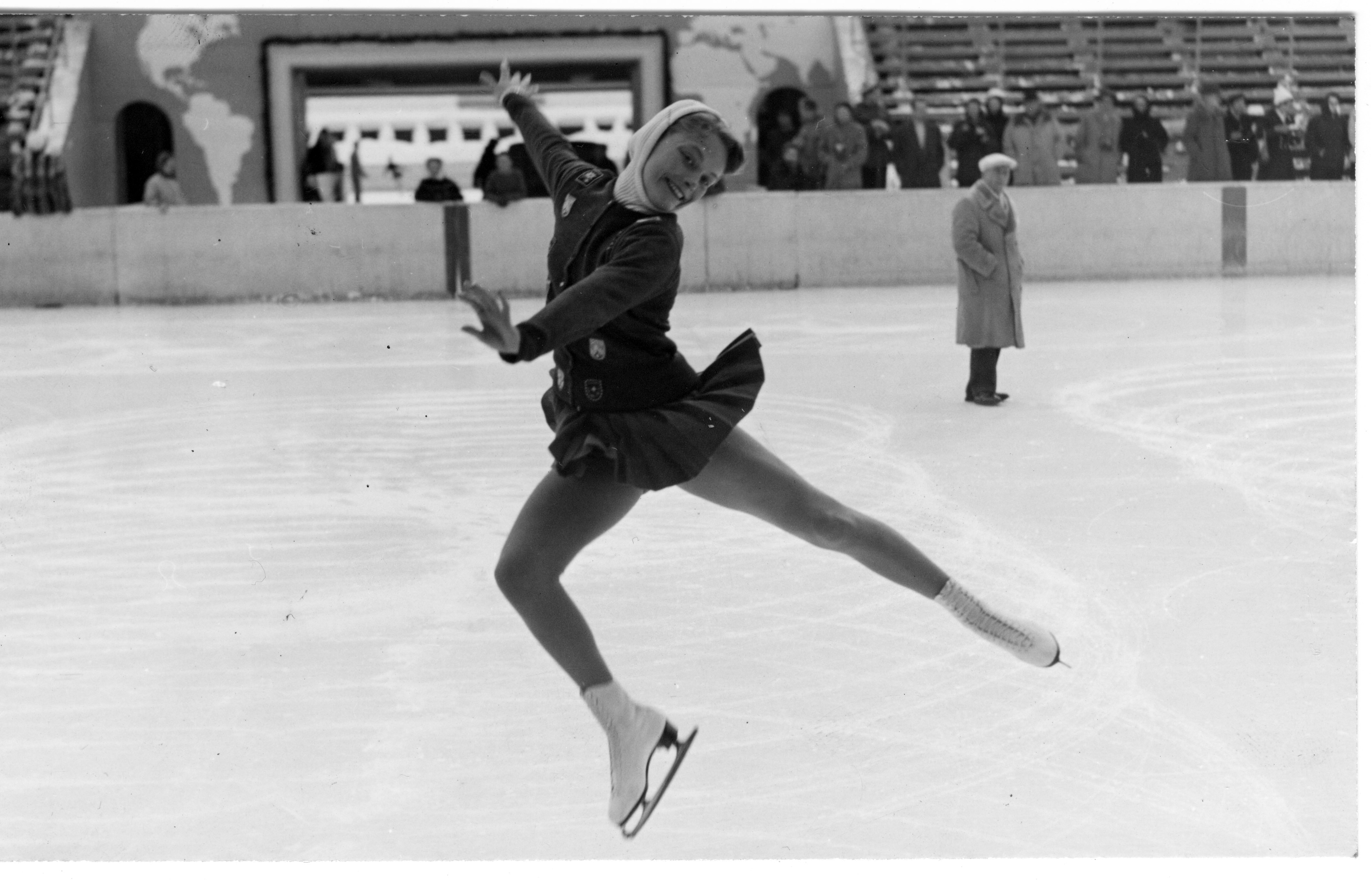 Carol Heiss performs a mid-air arabesque in a black and white photo in an indoor ice rink,