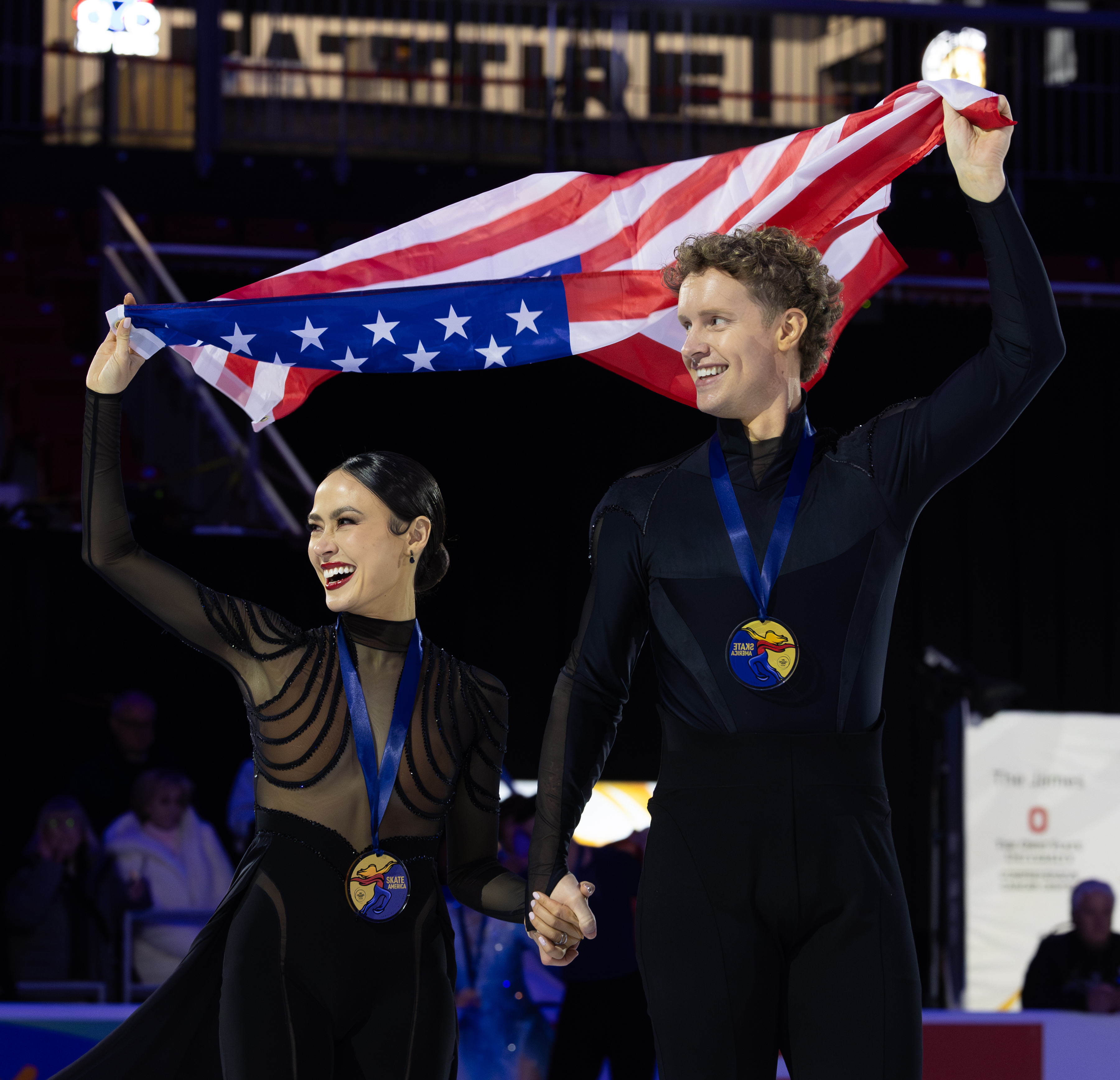Madison Chock (left) and Evan Bates (right) smile toward the crowd during the victory lap of the medal ceremony at 2025 Saatva Skate America. They both wear black, holding up the U.S. flab behind them, Skate America gold medals around their necks.