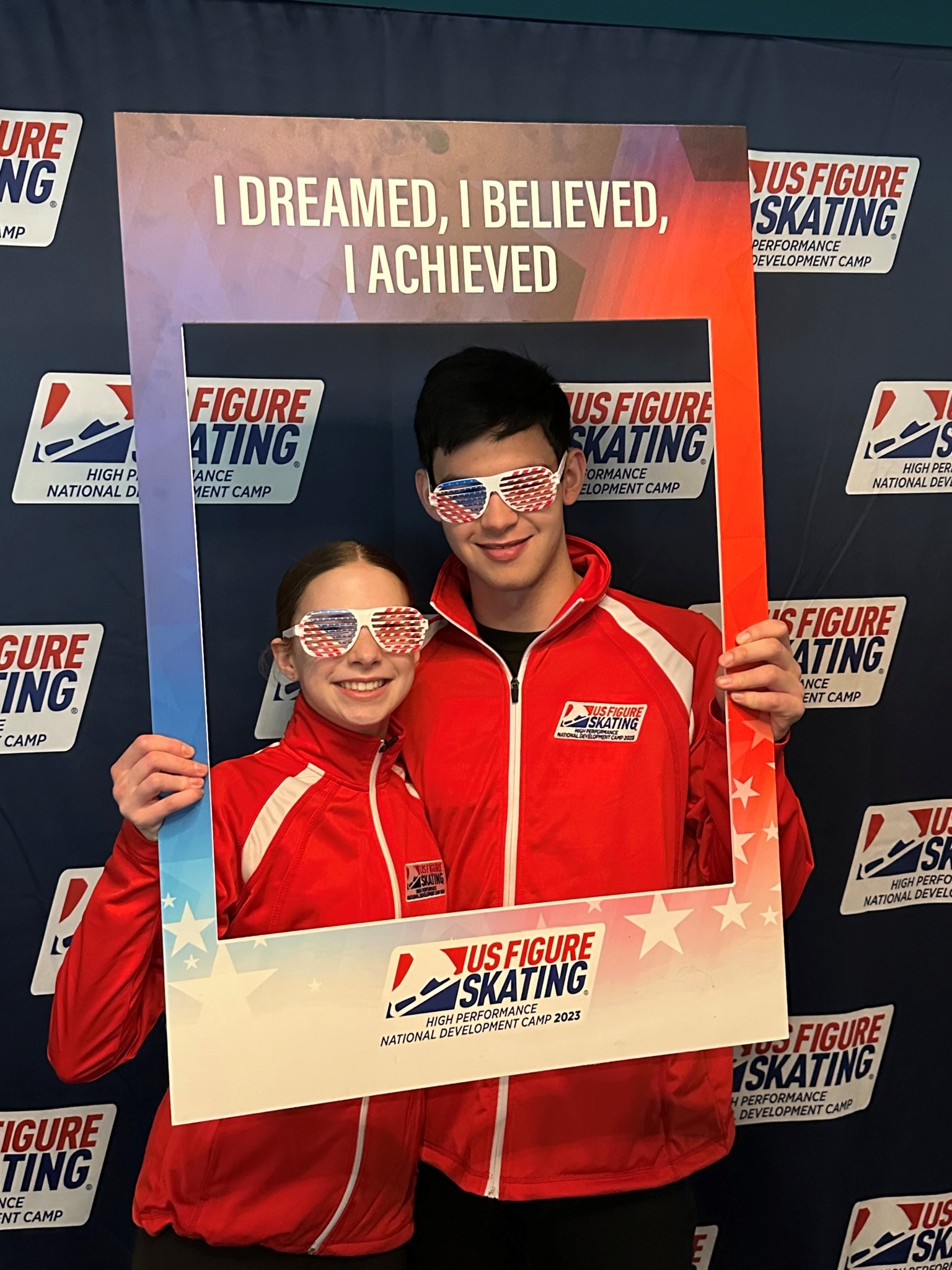 Clara Finkelstein and Mark Williams pose with a picture frame that says "I dreamed, I believed, I achieved" and funny red white and blue sunglasses Clara is a young white girl with brown hair tied back and Mark is a tall white boy with shirt black hair. They are both wearing red National Development Team jackets