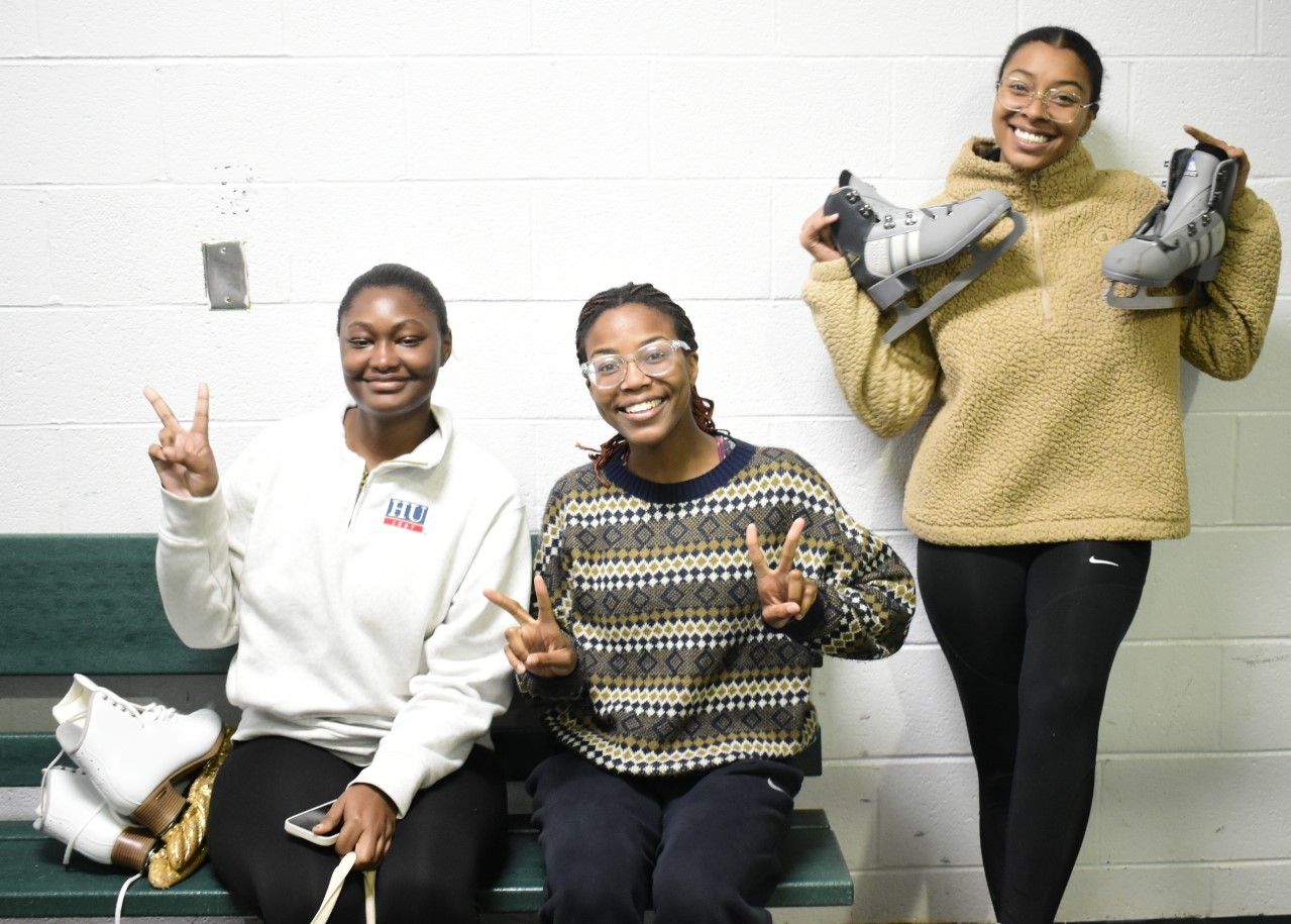 Three Black women smile for a photo as they put their skates on before getting on the ice. 