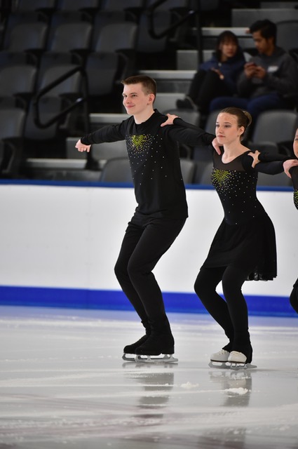 Tim performs with his synchronized skating team, the Rinkettes. Tim is a white teenager with short brown hair. He is wearing a long sleeve black shirt with gold detailing on the front. He holds on to the shoulder of another member of his team to the right of him