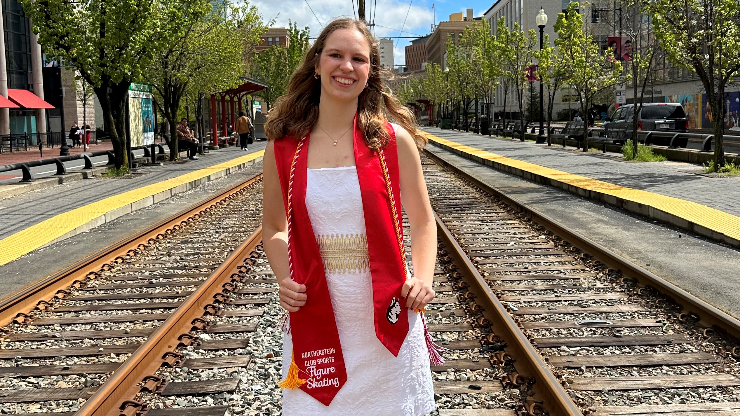 Elizabeth Klemm poses for a photo on train tracks. She is a young white woman with shoulder length blonde hair. She is wearing a white dress with a red grauation stole and a red and white graduation cord