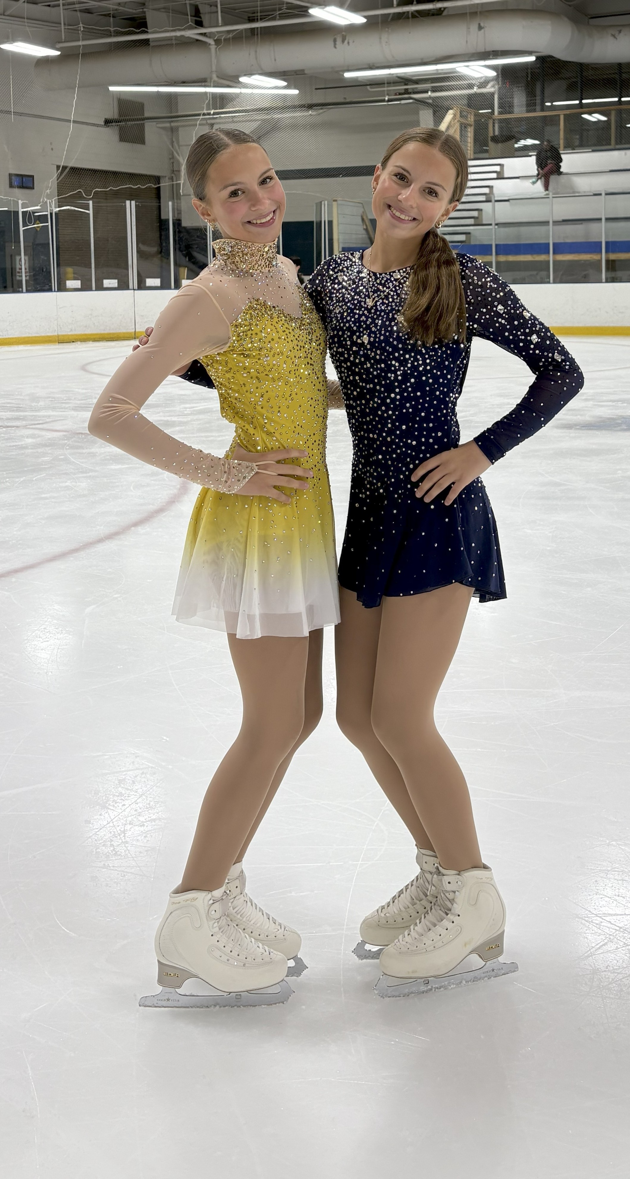 Ellie Flaks, dressed in a yellow skating dress, and Viv Flaks, wearing a purple skating dress, stand next to each other on the ice.
