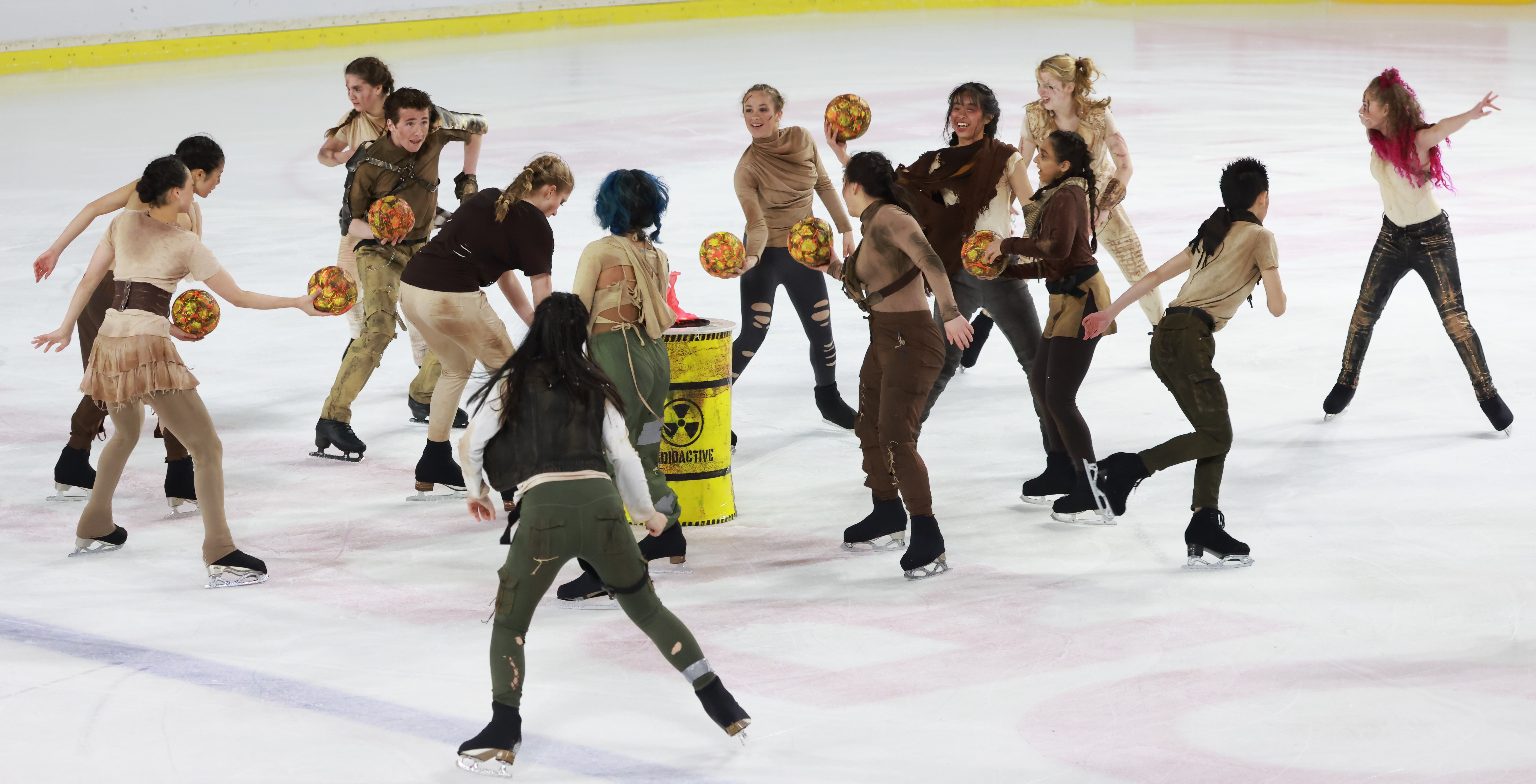 The team stands in a circle wearing tattered brown and tan clothing. They throw yellow balls into a yellow barrel with the word "radioactive" on the front