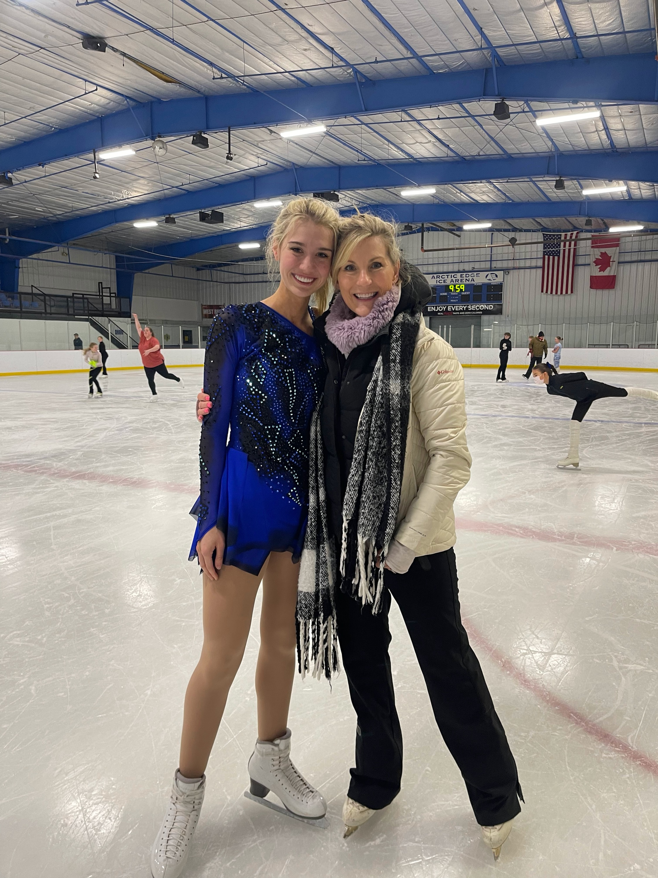 Elizabeth and her coach pose for a photo on the ice. Elizabeth (left) is a young white woman with long blonde hair wearing a long sleeve black and blue skating costume. Her coach (right) is an older blonde white woman wearing a beige winter coat and pink scarf. 