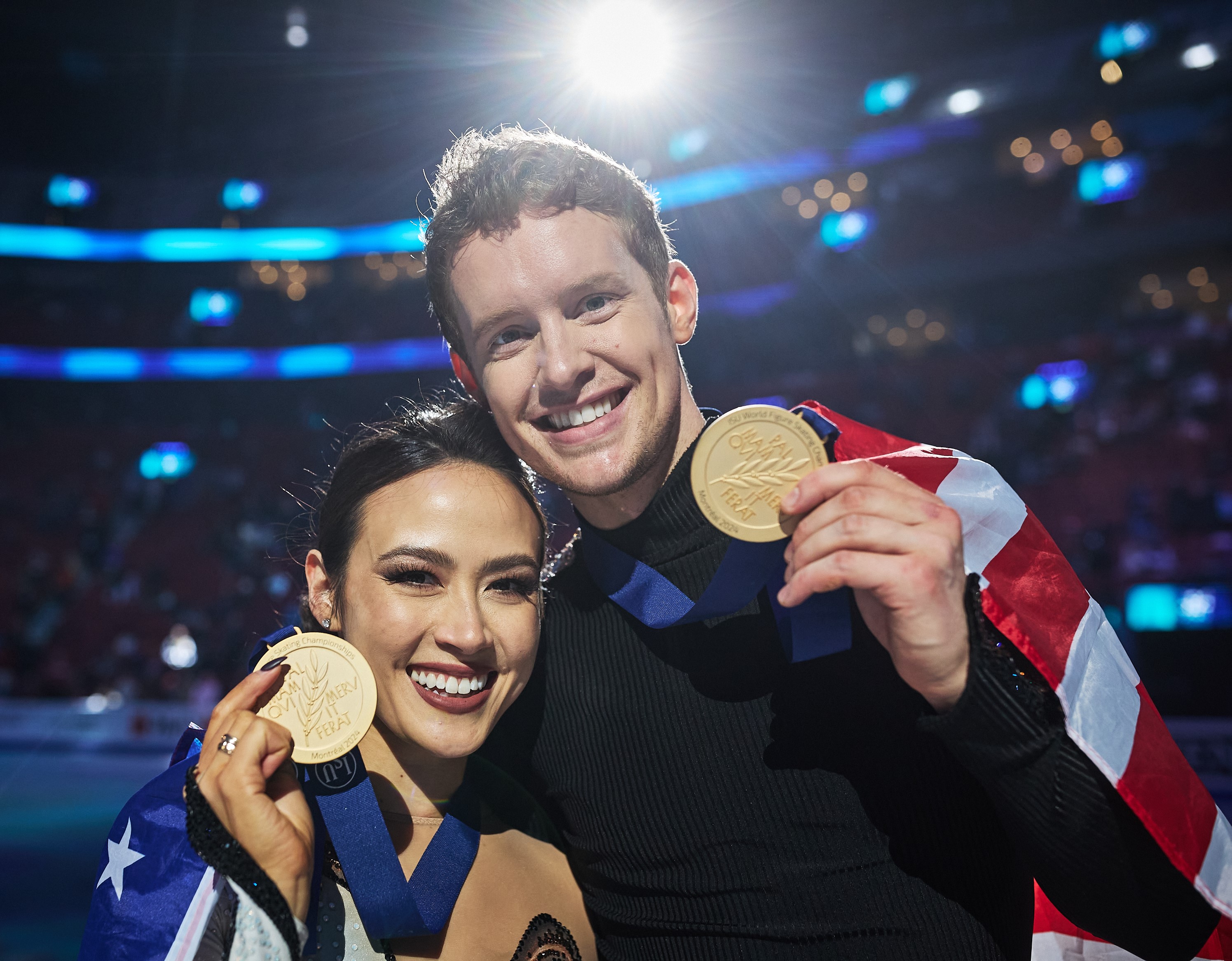 Madison Chock and Evan Bates, in a closeup shot, hold up their gold medals, the USA flag wrapped around them.