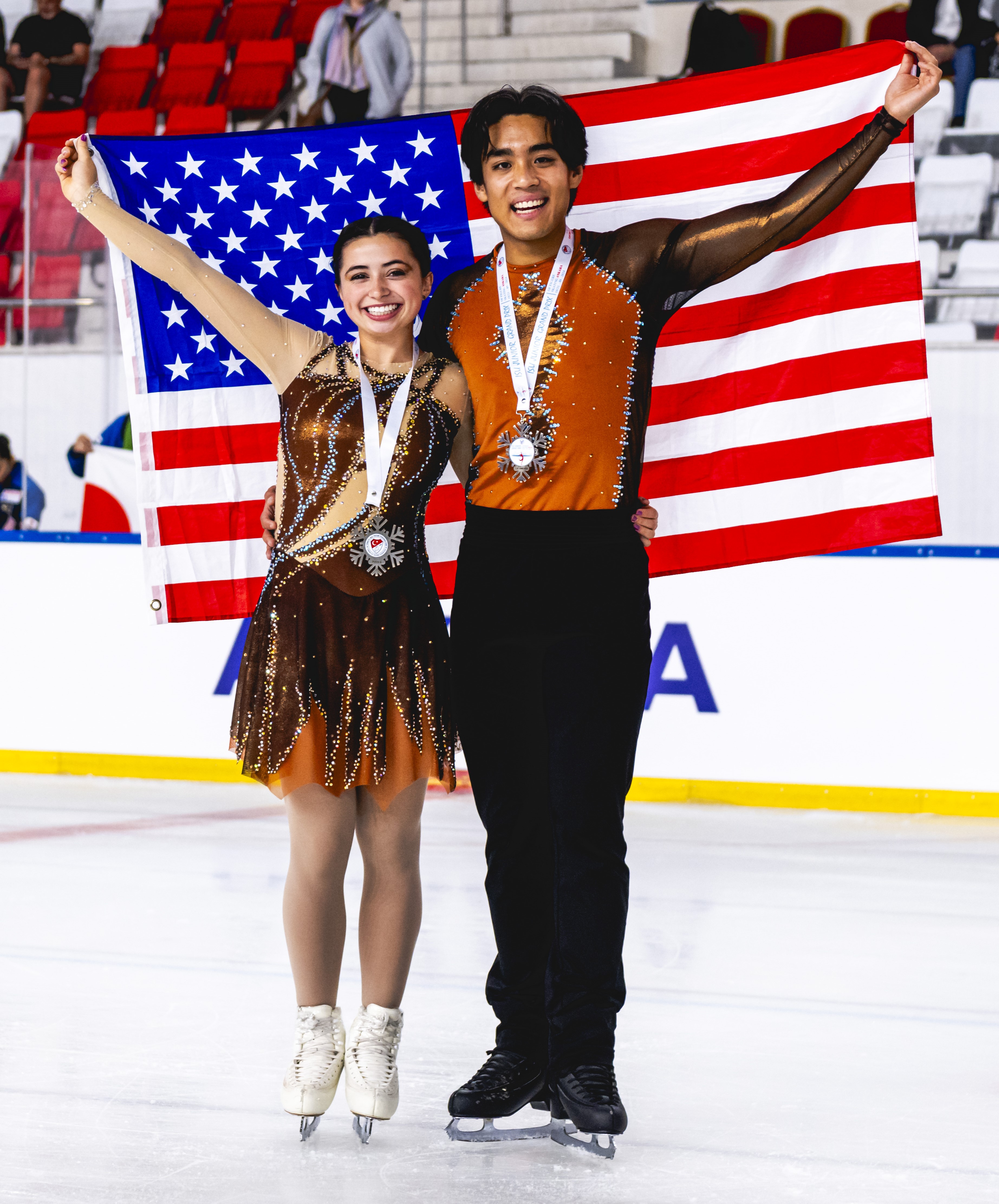 Olivia Flores and Luke Wang hold the U.S. flag behind them on the ice, smiling with silver medals around their necks. She wears a brown dress with jewels while he wears black pants with an orange and brown shirt.