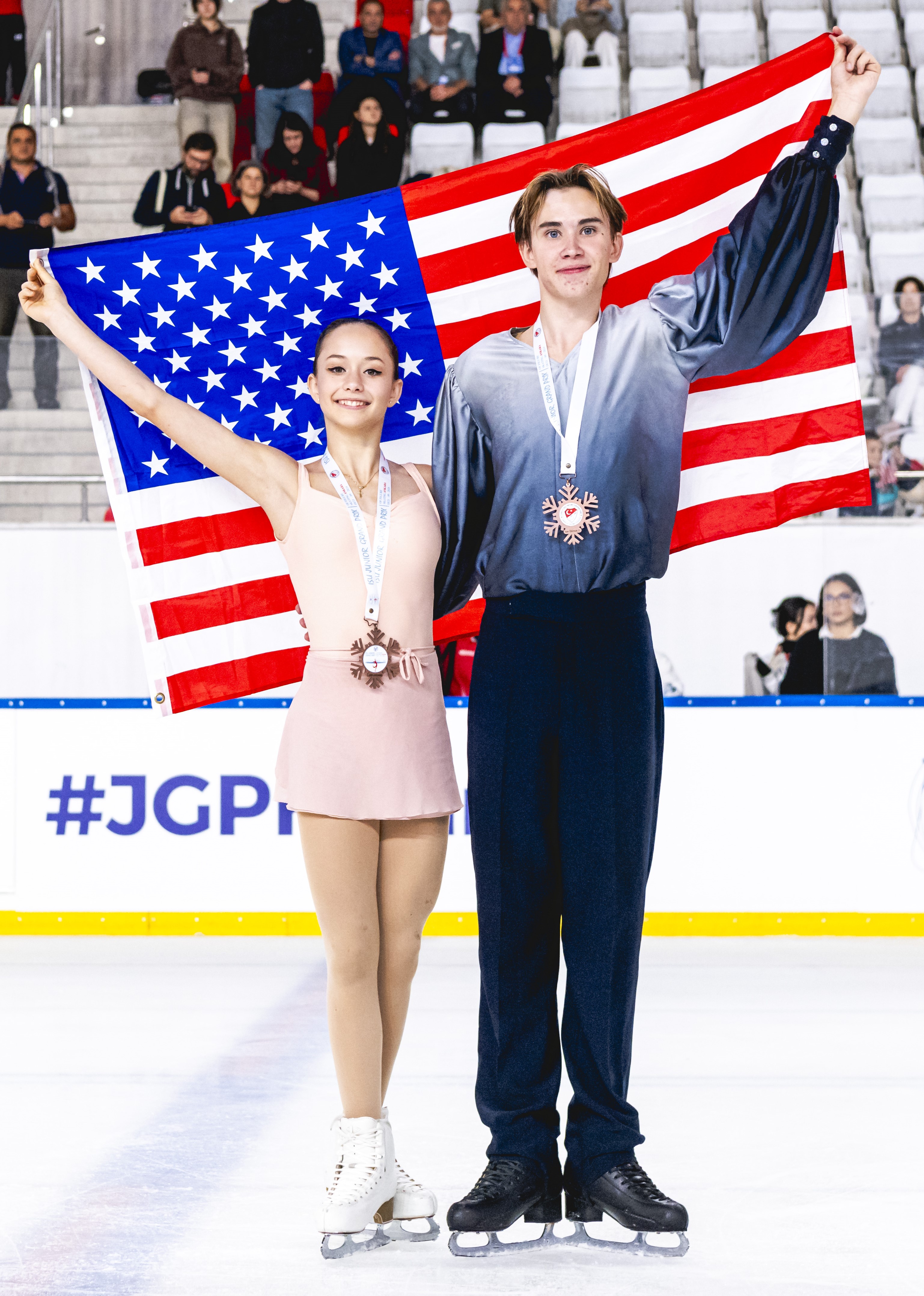 Hana Maria Aboian and Daniil Veselukhin hold the U.S. flag behind them on the ice, smiling with bronze medals around their necks. She wears a light pink dress while he wears an ombre white to navy shirt with navy pants.