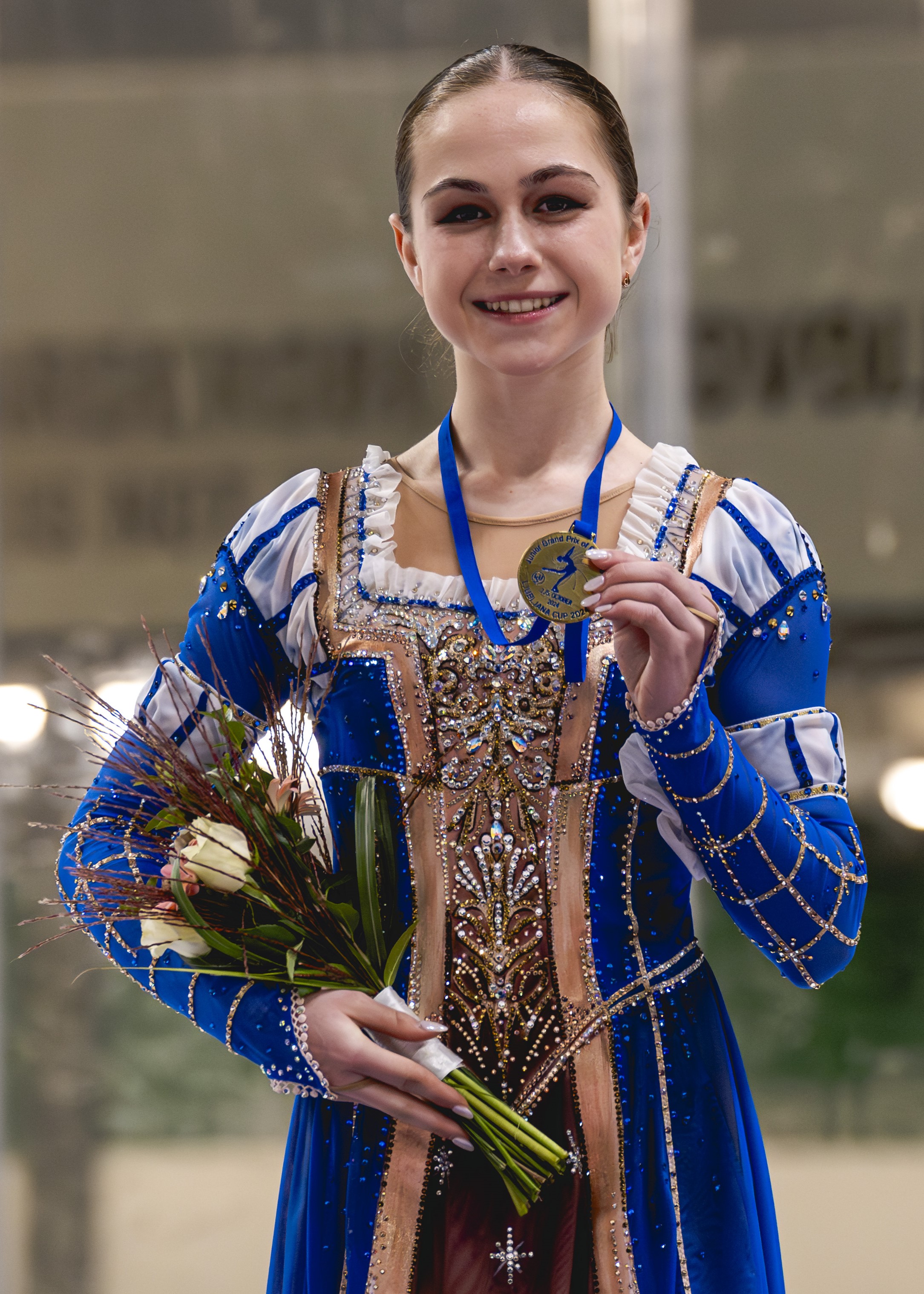 Sophie Joline von Felten smiles while standing on the podium at JGP Slovenia. He holds her medal in one hand and flowers in the other. She is wearing a blue skating costume inspired by Romeo and Juliet