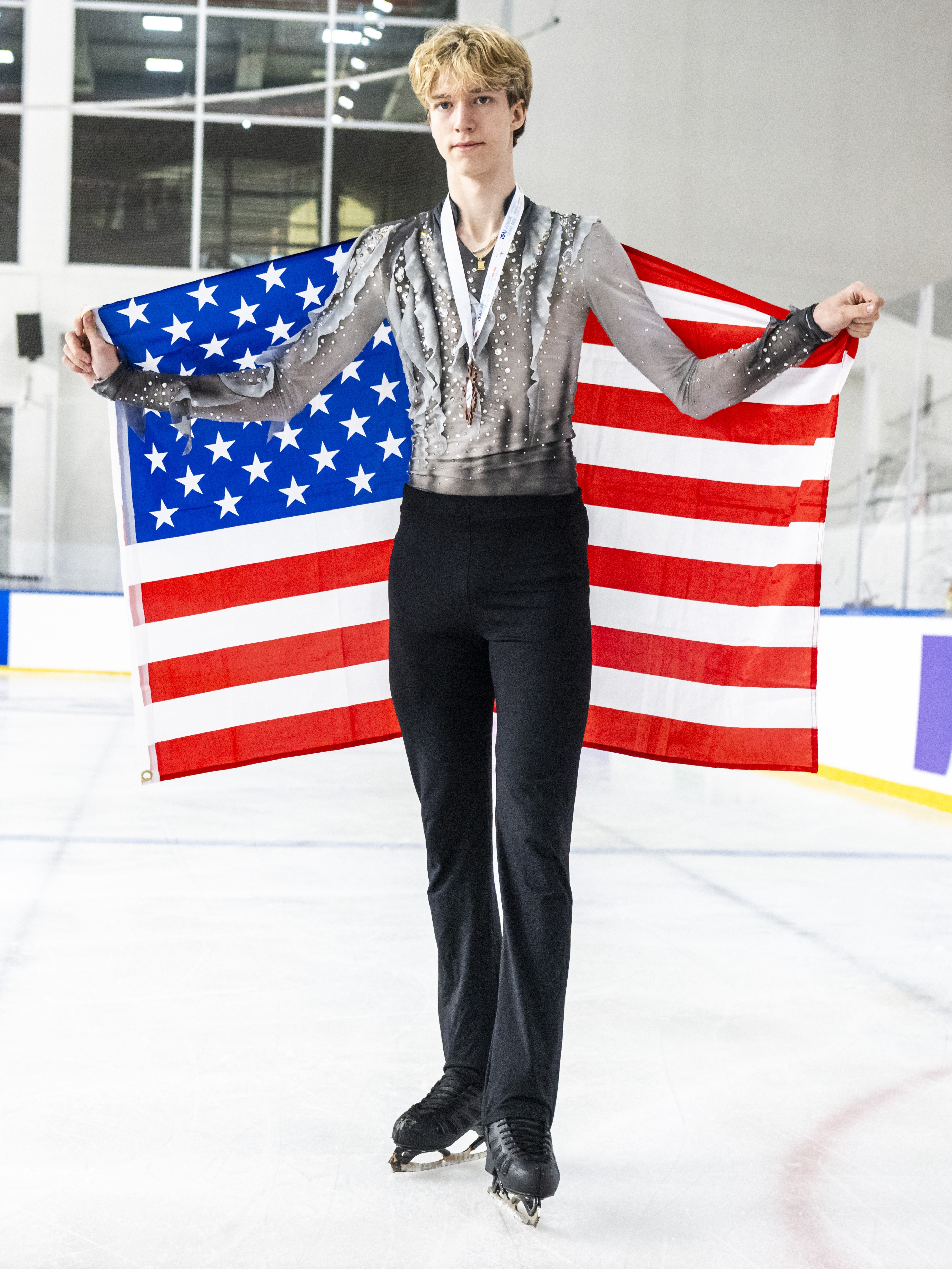 Lucius Kazanecki stands holding an American flag behind him with a bronze medal with a white ribbon around his neck. Lucius is a young man with blonde hair wearing a long sleeve black and tan top with jewel detailing, black pants and black skates.
