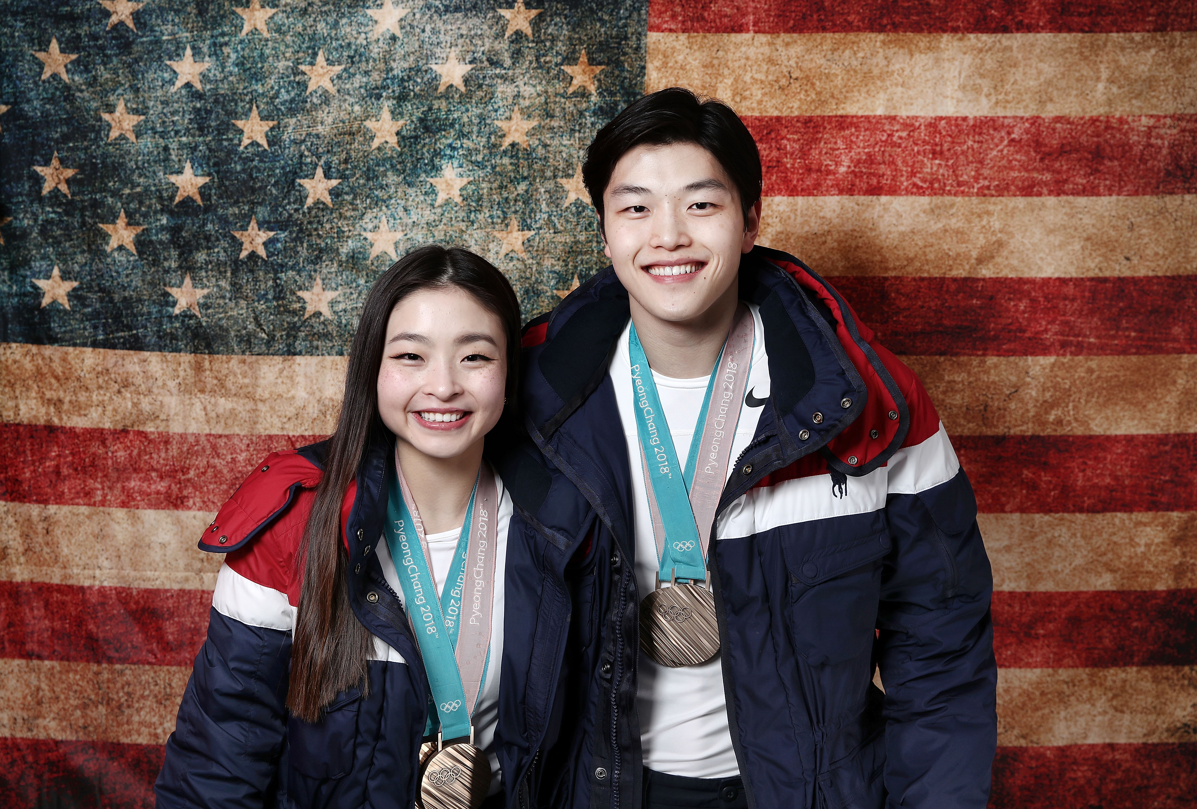 Maia and Alex Shibutani wearing their Olympic medals in front of a U.S. flag backdrop.