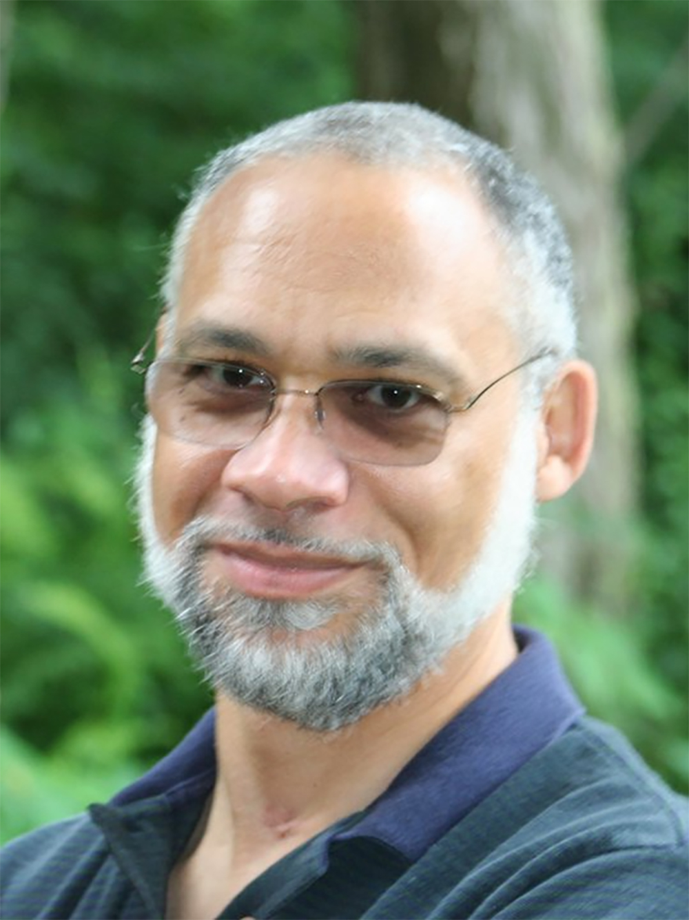 Headshot of Alan Mathews. e is a middle aged African American man with grey short hair and a beard. He wears glasses and a blue and green collared shirt