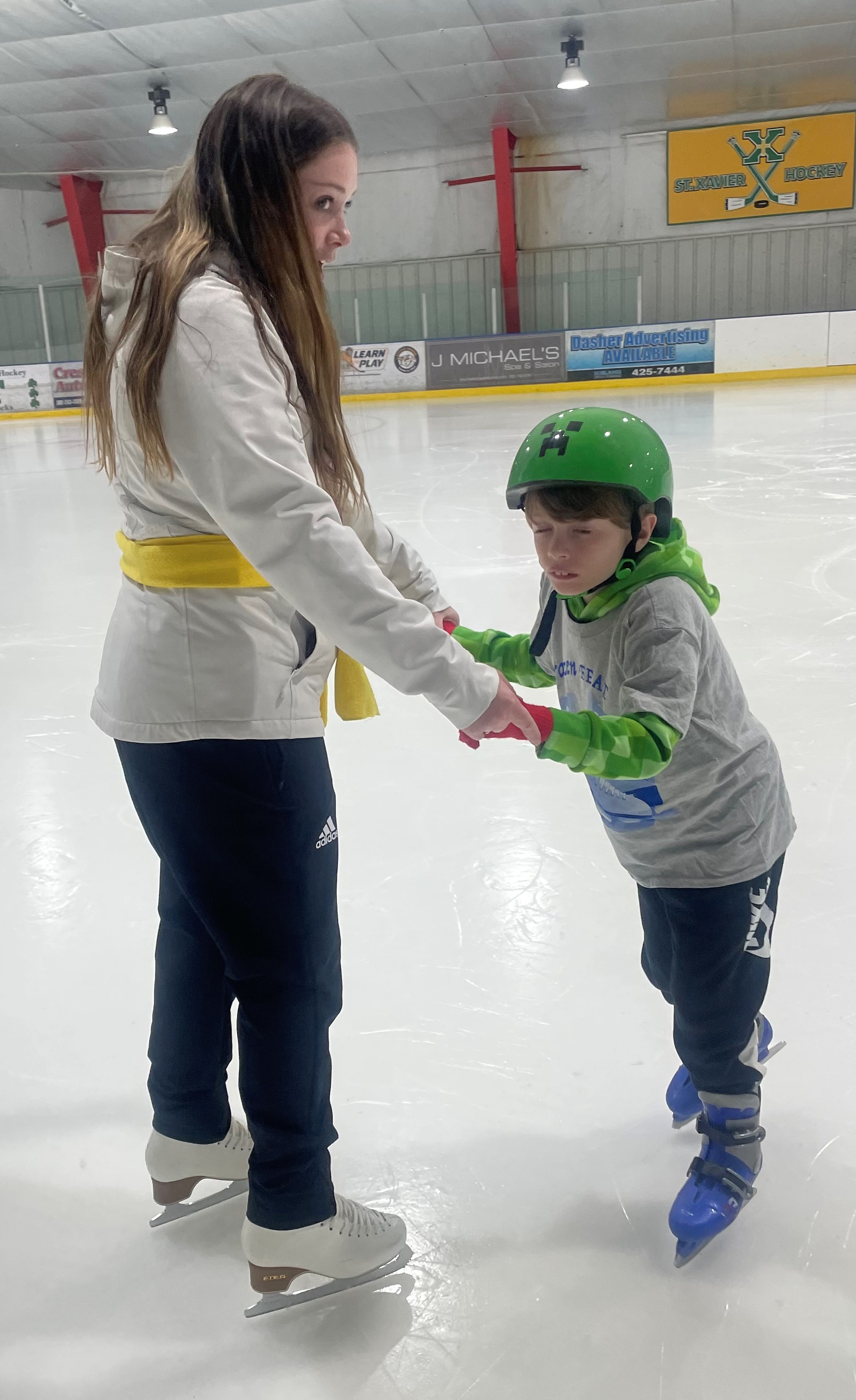 A young girl helps a young boy with autism skate. The girl (left) has long brown hair and is wearing a grey sweatshirt and black pants. The young boy (left) is wearing a bright green Minecraft helmet, grey tshirt over a green hoodie, and black pants. The girl is holding both of the boy's hands to help him balance on the ice 