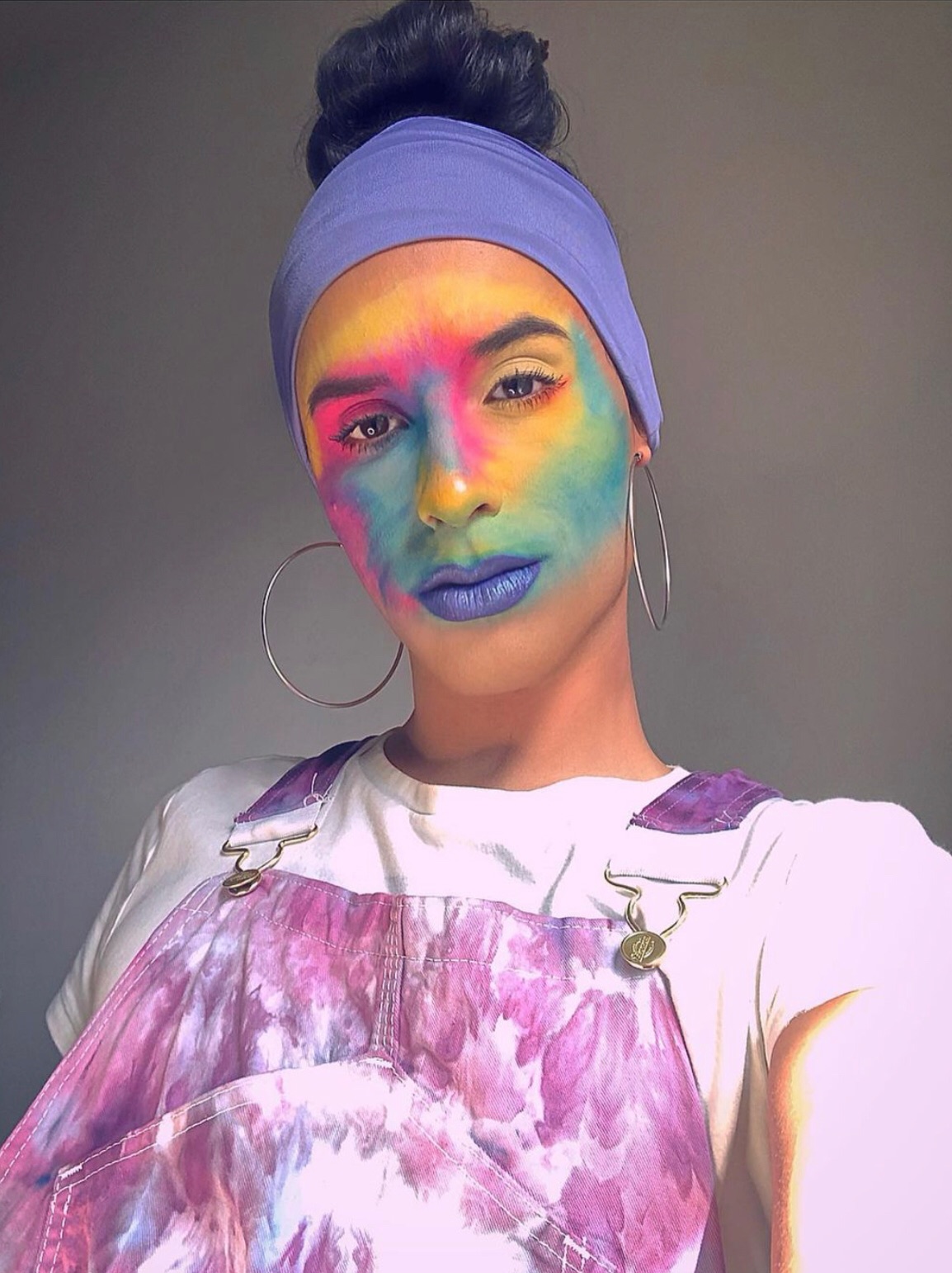 A headshot of Eliot Halverson with her brown hair in a bun and face painted with rainbow colors.