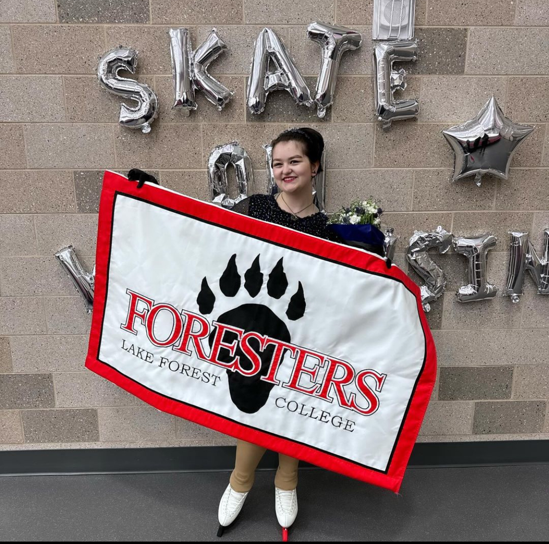 Tobi Ellis stands in front of a wall of silver balloons that spell out "Skate on Wisconsin" holding a sign that says "Foresters" in red letters, and below it in black it says "Lake Forest College" The flag has a white background, red border and features an animal footpring behind the text. Tobi has their brown hair tied up and they are wearing a black skating costume and skates. In their right hand is a bouquet of flowers. 