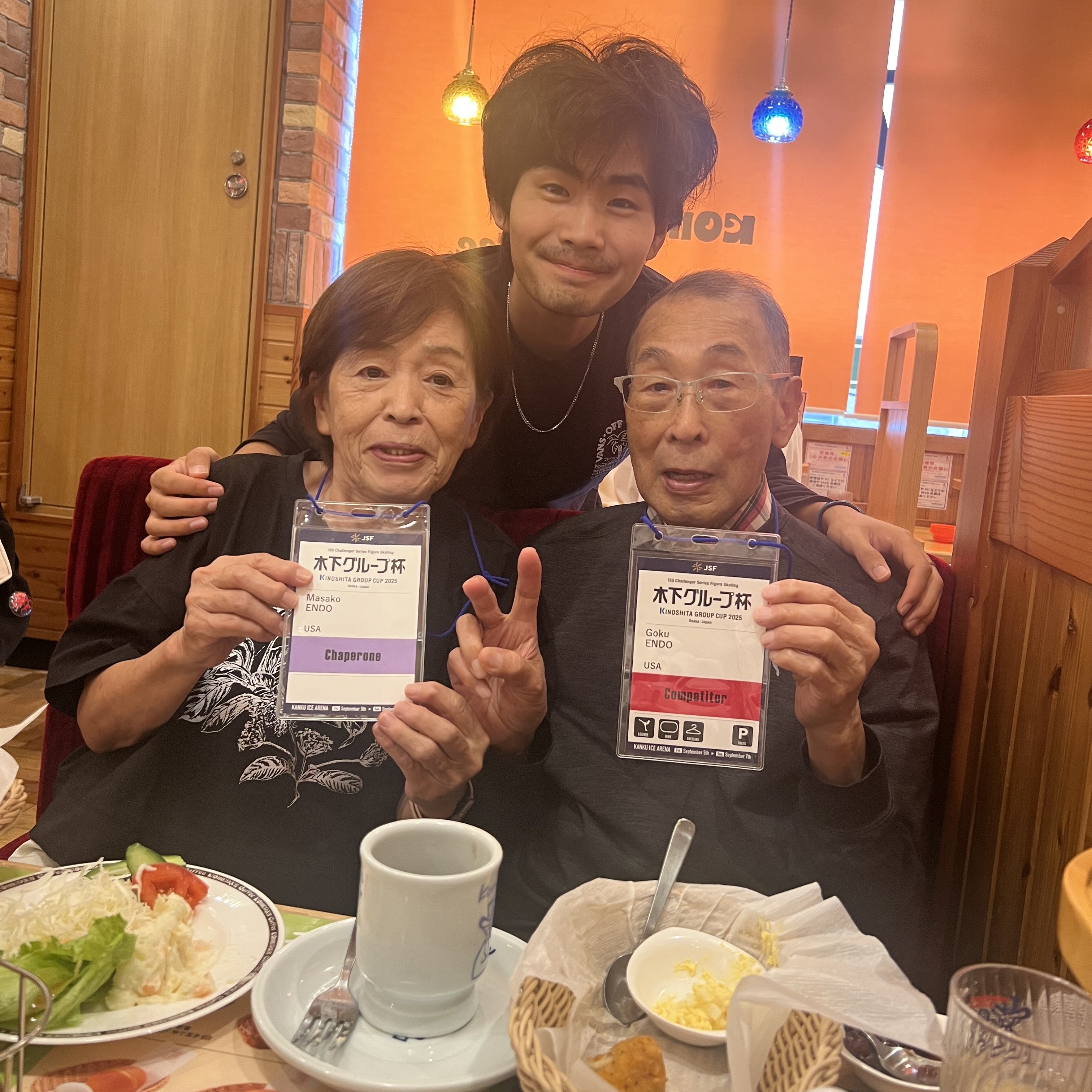 Goku stands behind his grandparents as they pose with their credentials from the Challenger Series event with Japan. They are at a restaurant.