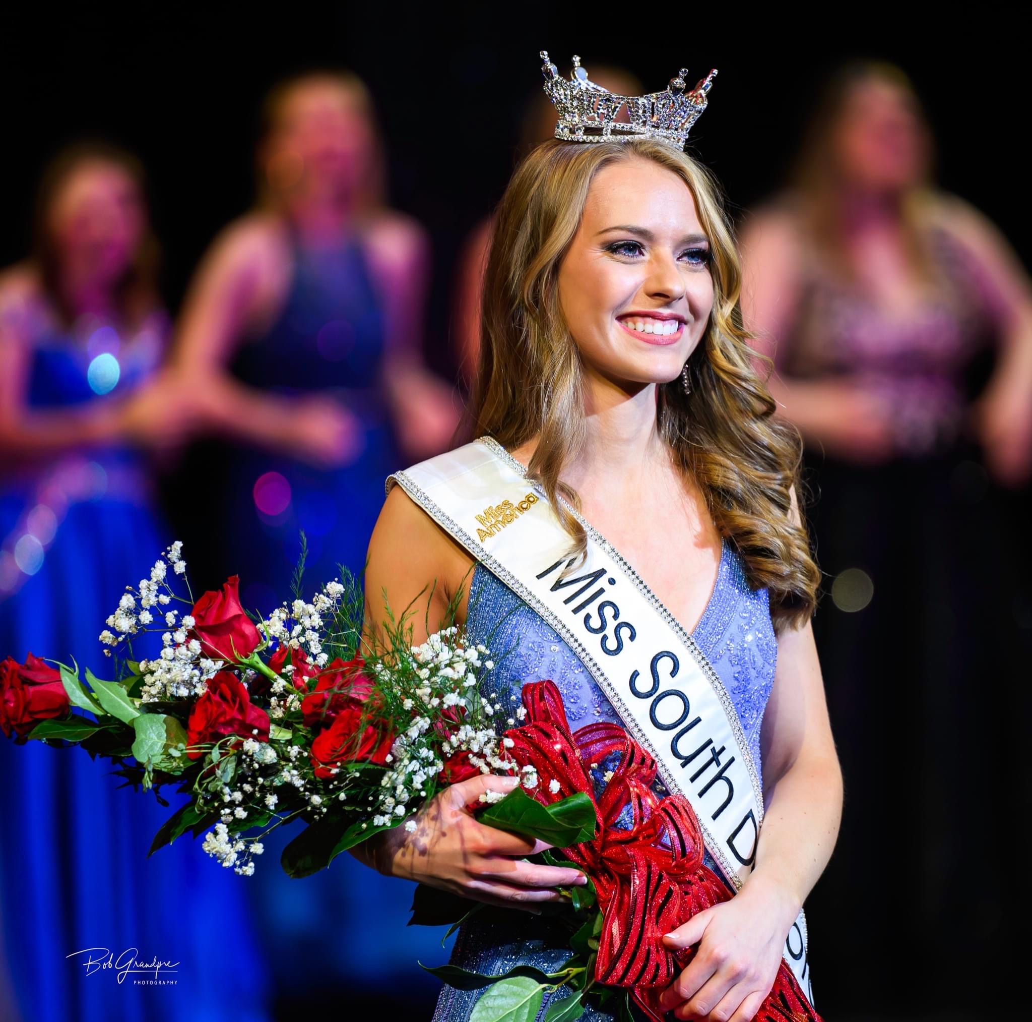 Hunter Widvey in a light blue dress with the Miss South Dakota sash holding flowers with a crowd on her head.