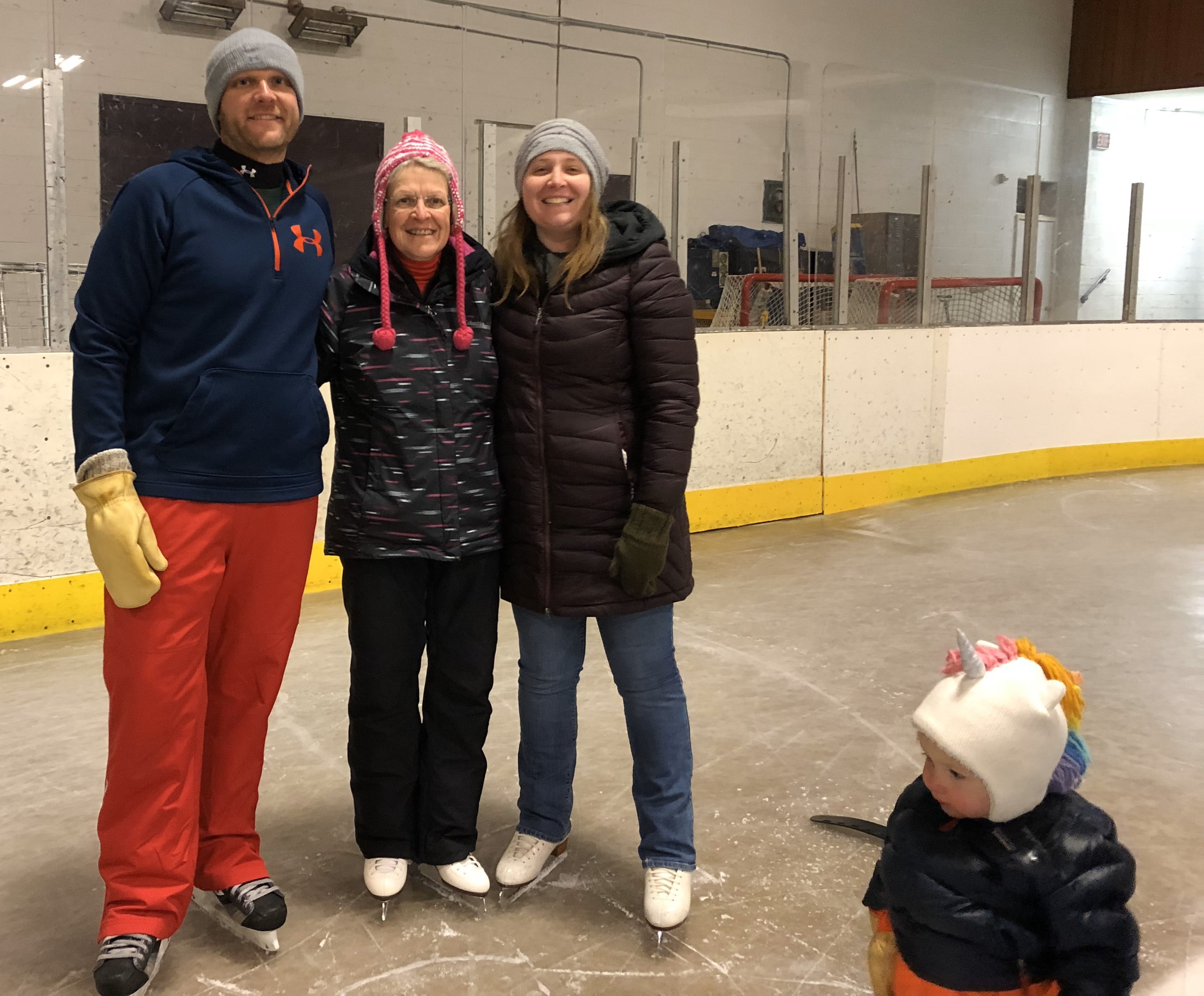 Linda poses with her son and daughter at Babbitt Arena. They are all wearing winter attire and knit hats. A young girl wearing a knit unicorn hat sits in the foreground.