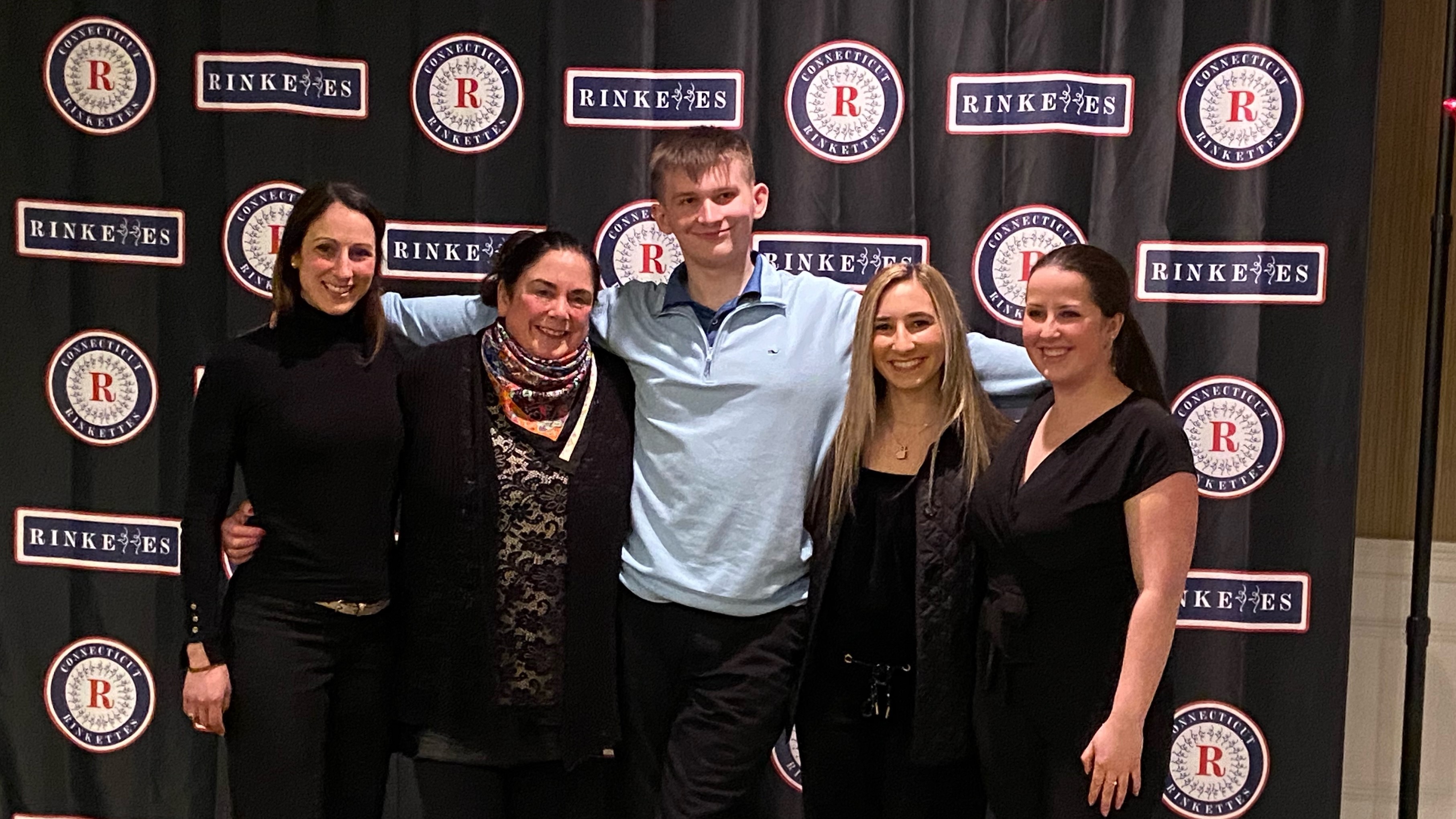 Tim poses in front of a Rinkettes backdrop with some of his coaches. Tim (center) is a tall Caucasian teenager with short brown hair. He is wearing a light blue quarter zip and black pants. His coaches standing around him are all older women wearing black clothing