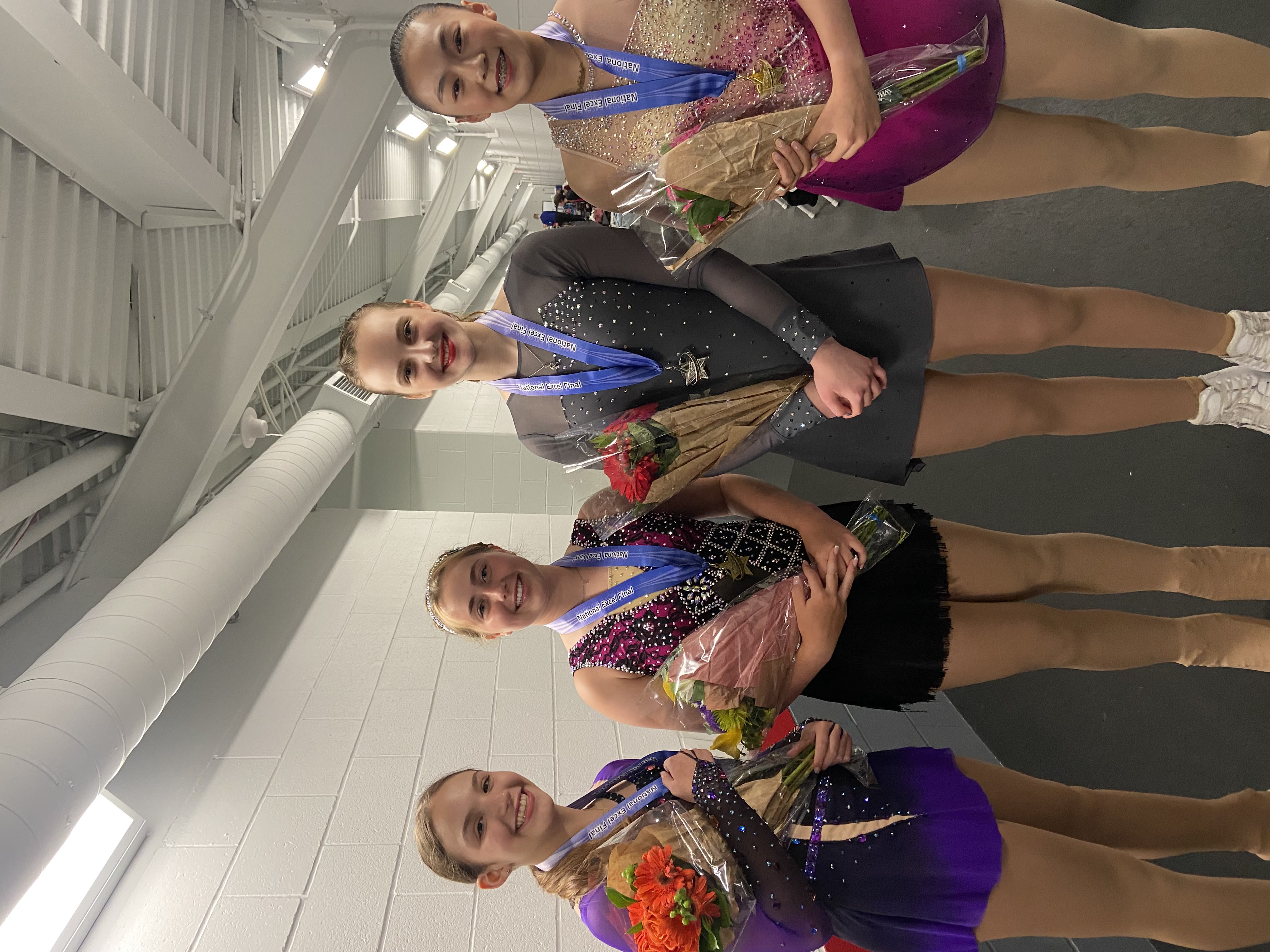 Junior women in their skating costumes hold flowers while in their skates in a rink hallway.