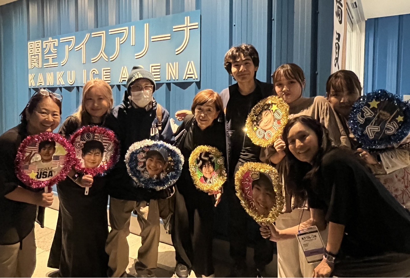 Goku poses with his family and friends, who are all holding sticks with pictures of him. They are in a hallway at the ice arena in Japan.