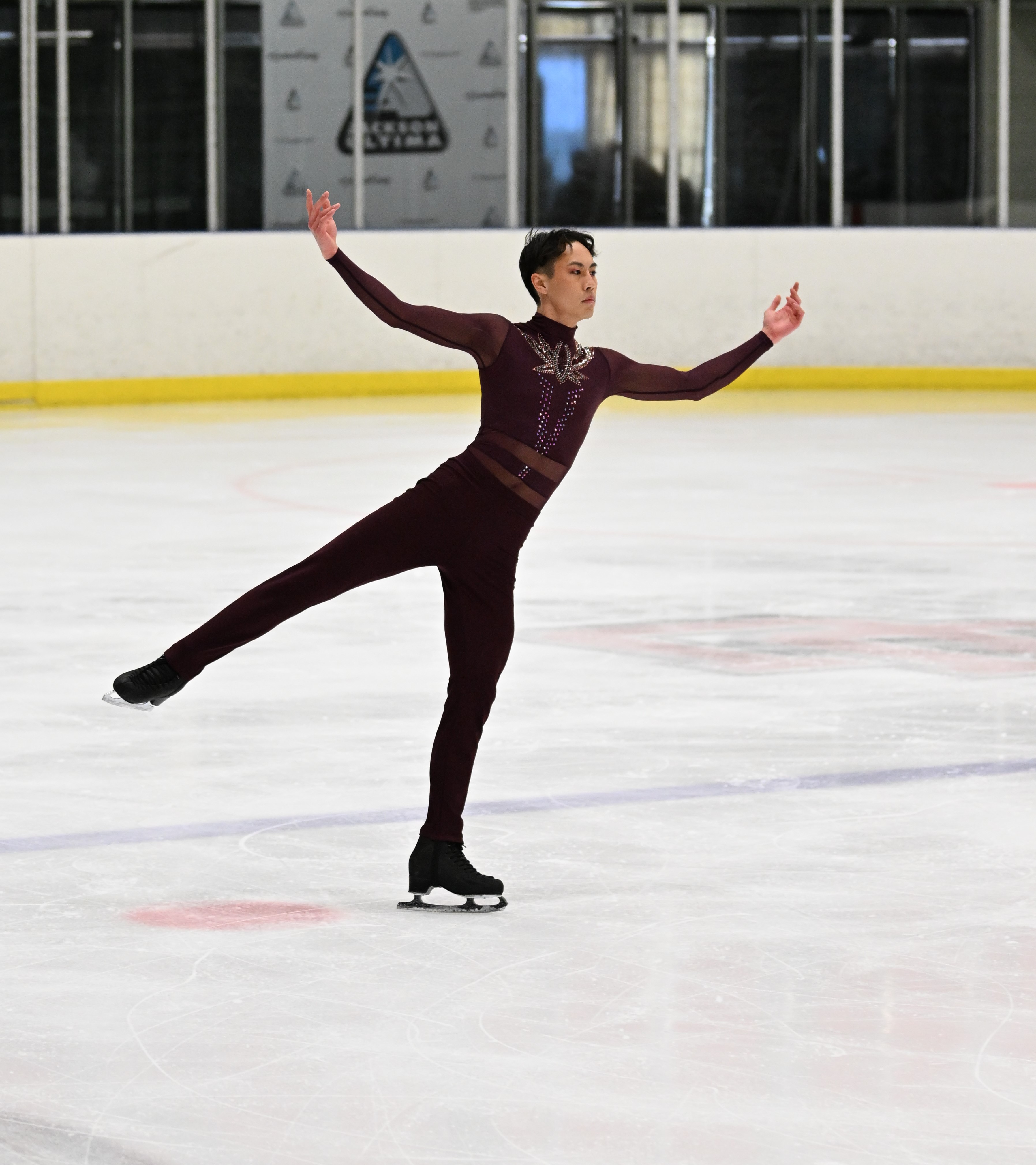 JY Sun competes at the 2023 U.S. Adult Figure Skating Championships. JY is an Asian Man with short black hair. He is wearing a long sleeve maroon top with jewels on the front and black pants. He stands with his arms outstretched and his right leg extended outward