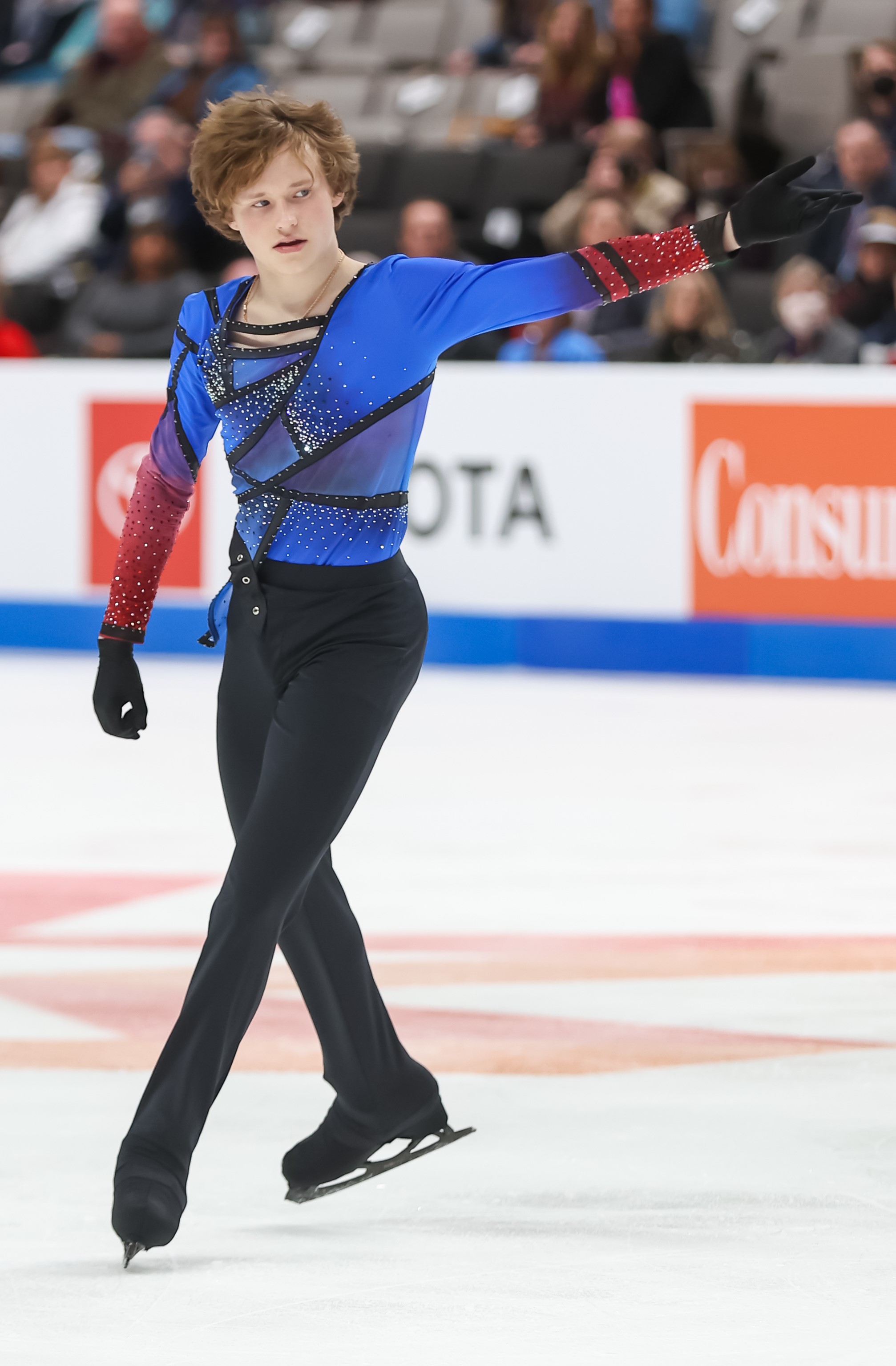 Ilia Malinin competes in the men's free skate at the 2023 Toyota U.S. Figure Skating Championships. He is wearing a blue long sleeve shirt with red sleeves. He holds his arm out to the right and looks off to the right