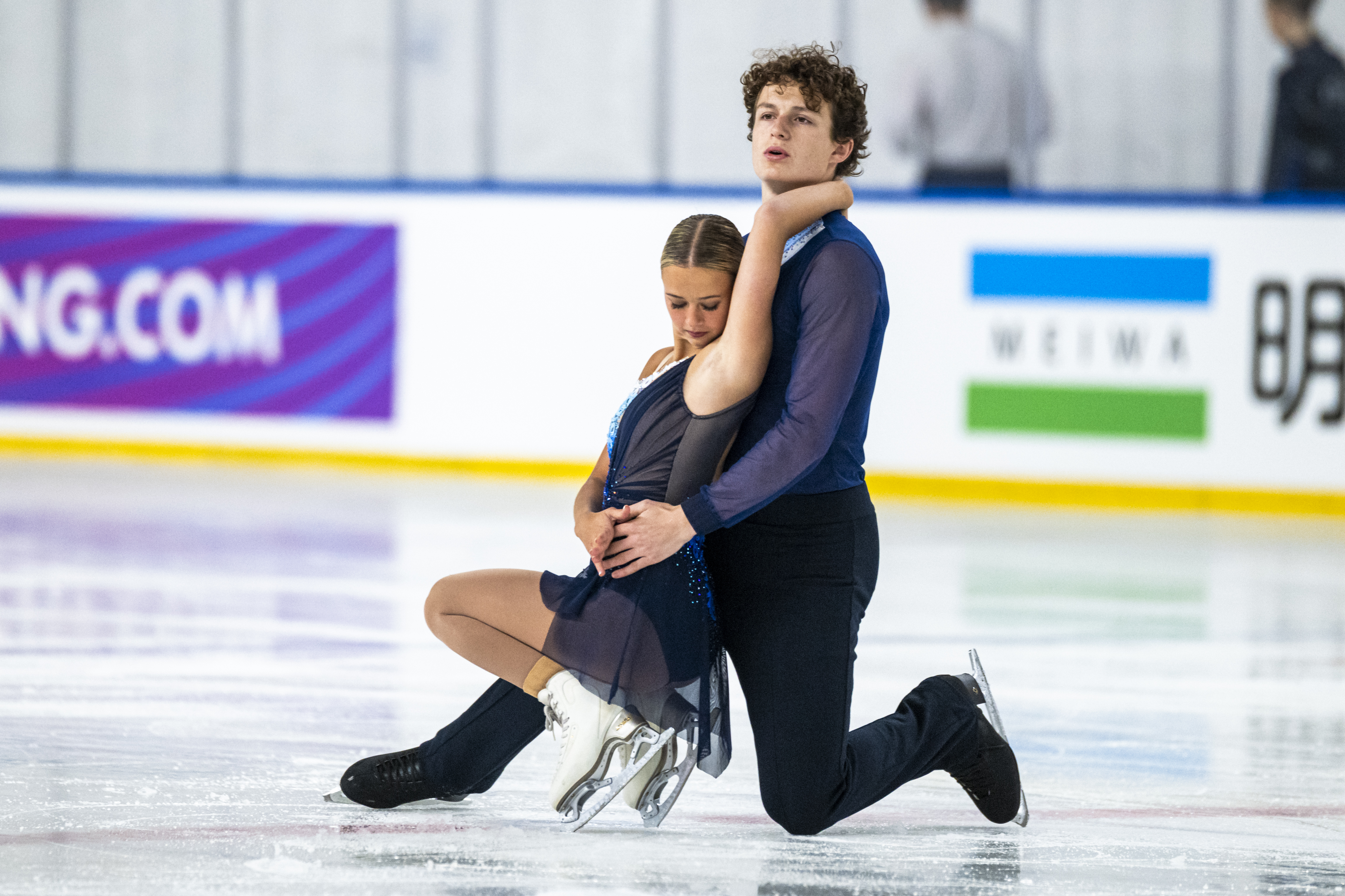 Luke Witkowski, kneeling on the ice, has his arms around partner Sofia Jarmoc who is facing away from him. Both are wearing violet-colored costumes.