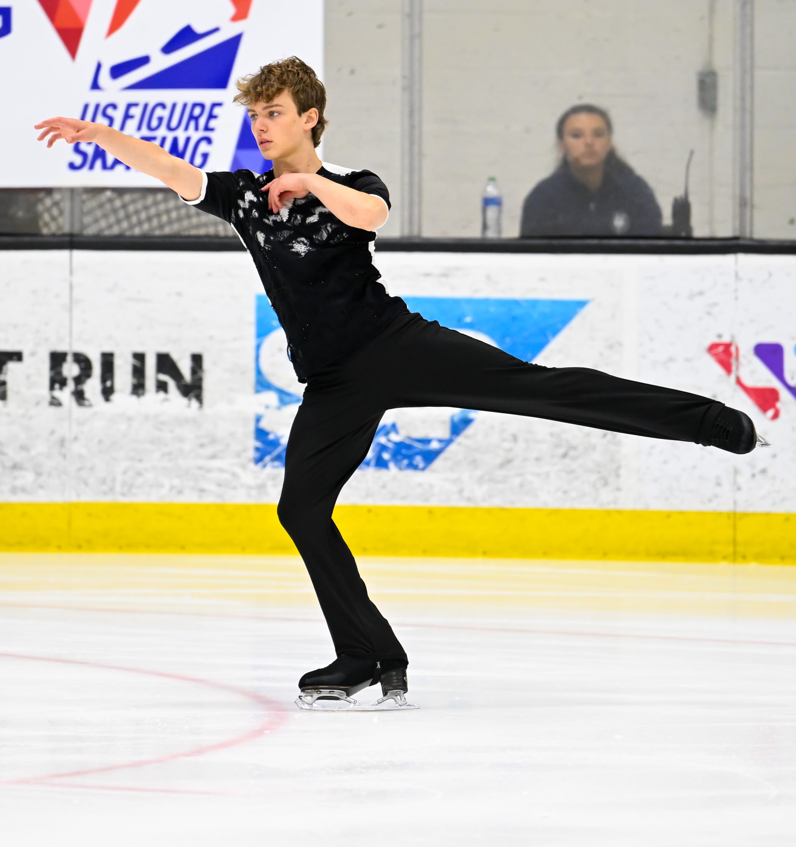 Andriy Kratyuk skates with one leg and one arm extended away from his body as he looks forward. He has short blonde hair and he is wearing a black short sleeve top with white trim and black pants