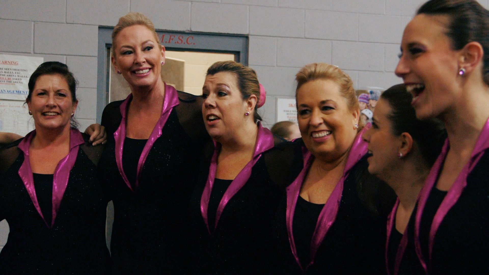A group of synchro skaters and laugh together off the ice with their arms around each other.