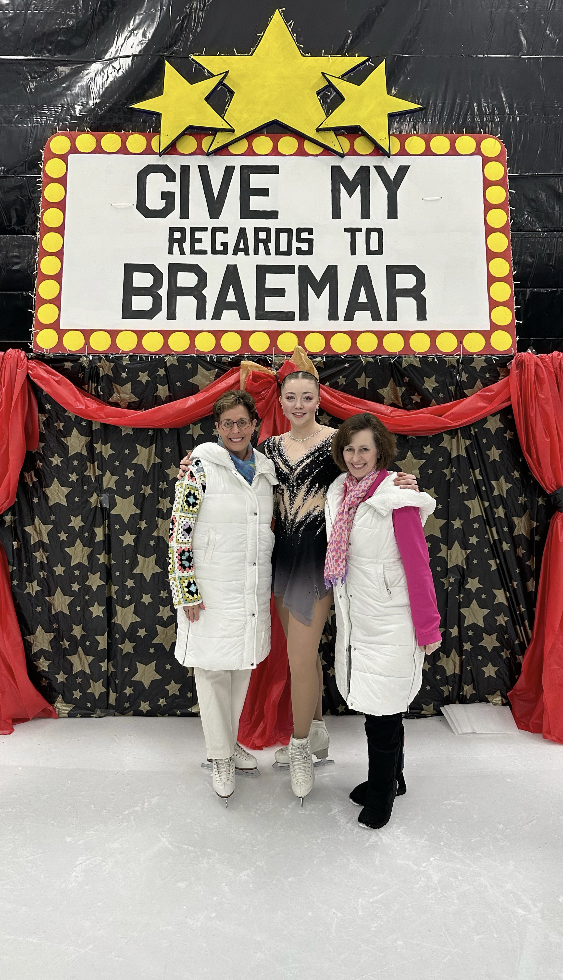 Skater Ava Thomas, middle, with Loni Keenan, left, and Kathleen Gazich in front of a tall sign that says: Give My Regards to Braemar
