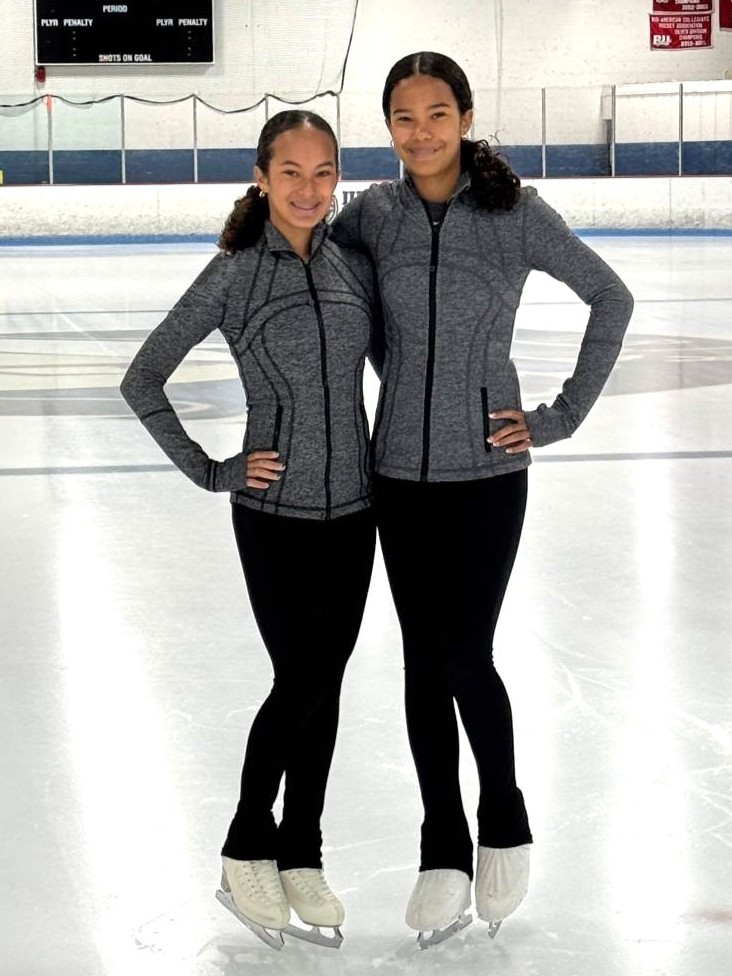 Mari Flores and Ellie Flores, both wearing gray tops and black pants, stand side by side on the ice.