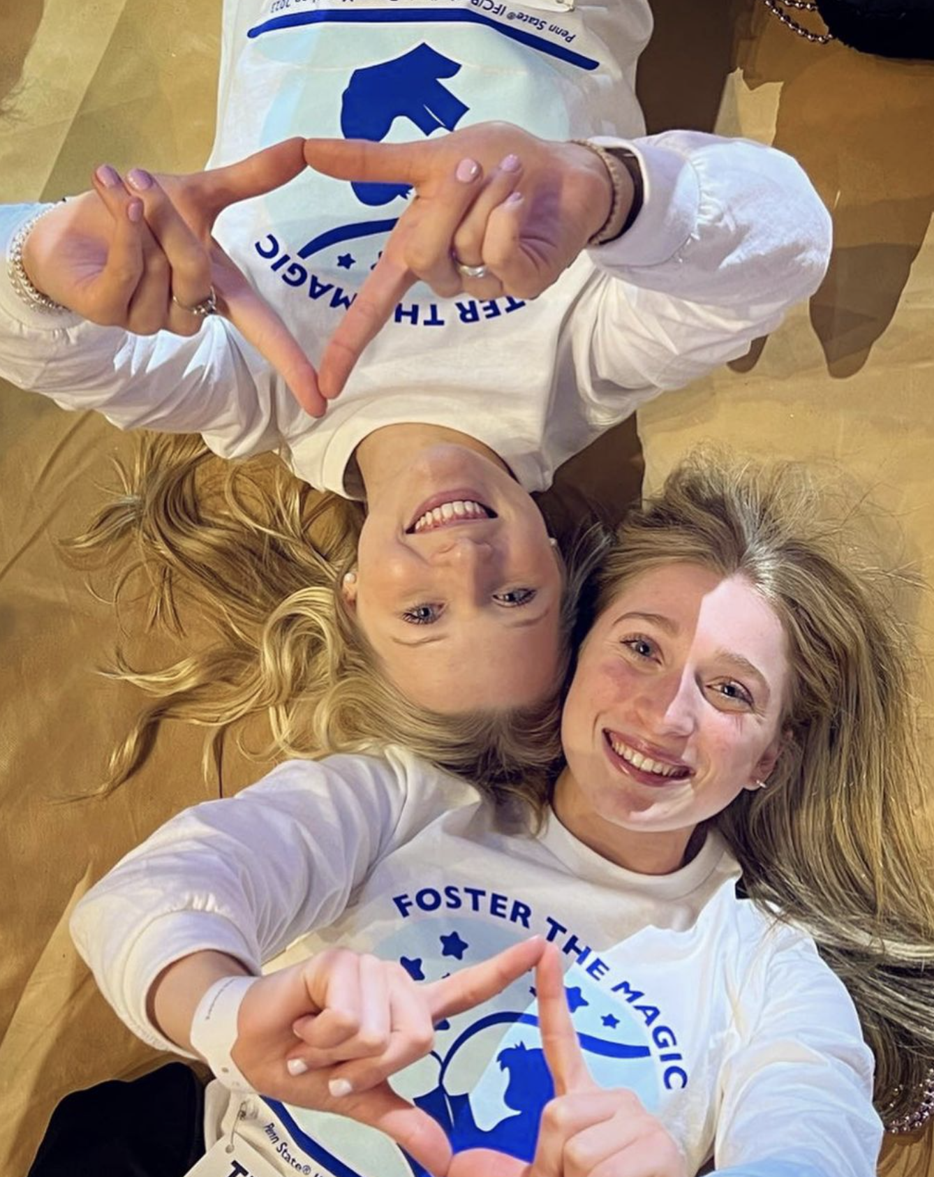 Marissa and Katie lay on the ground while getting ready for their 46 hour dance marathon. Both are yng Caucasian woman with long blonde hair. They hold up the official hand sign if the THON event by making a triangle with their pointer finger and thumb