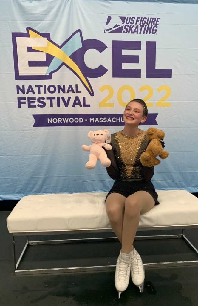 Photo of Chloe Taurel smiling. She is wearing a black sparkly figure skating costume and she is holding a stuffed animal in each hand