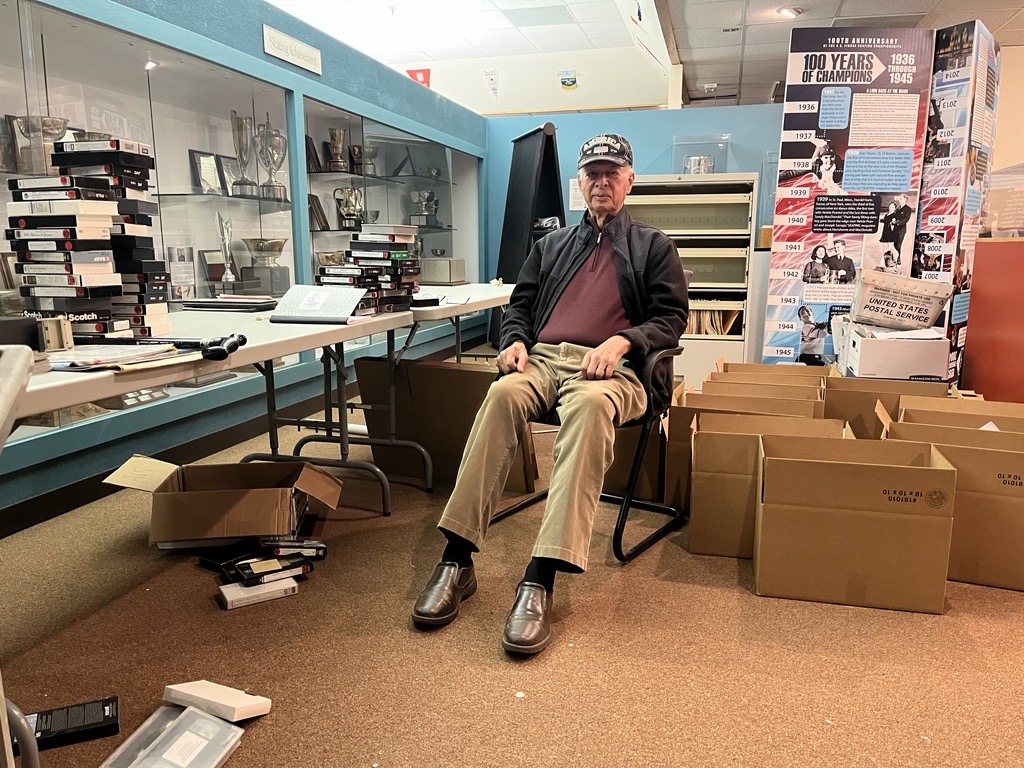 Photo of Bill volunteering at the World Figure Skating Museum and Hall of Fame. He is wearing a hat with an American Flag on it a dark red shirt, black leather jacket and khakis.