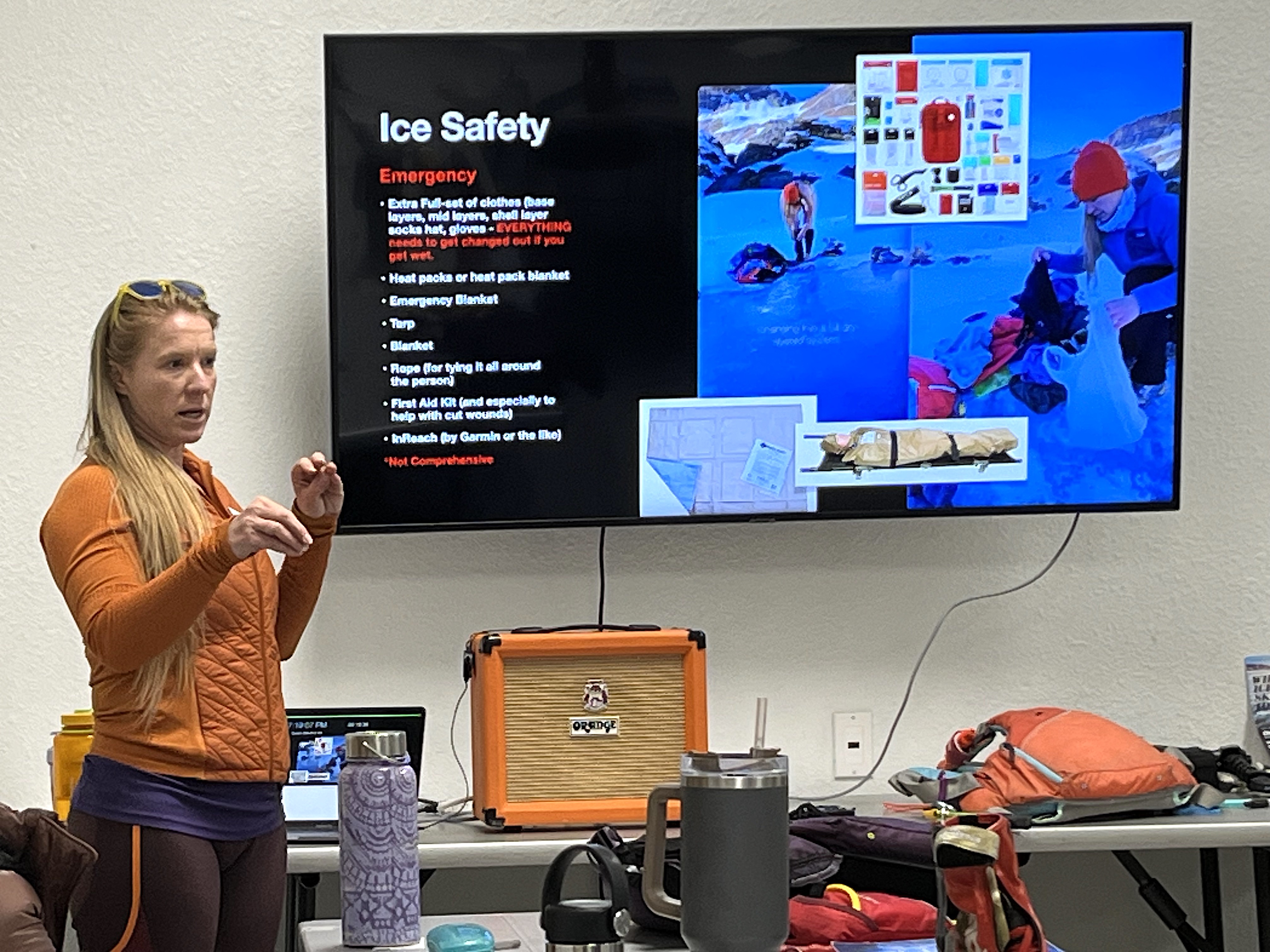 Laura stands in front of a tv talking to her students with Powerpoint slide titled Ice Safety on the screen. She has long blonde hair and is wearing a love sleeve orange shirt
