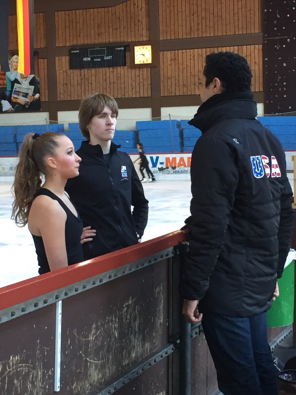 Nathan Truesdell, wearing a Team USA jacket, coaches from the boards as an ice dance team listens while on the ice.