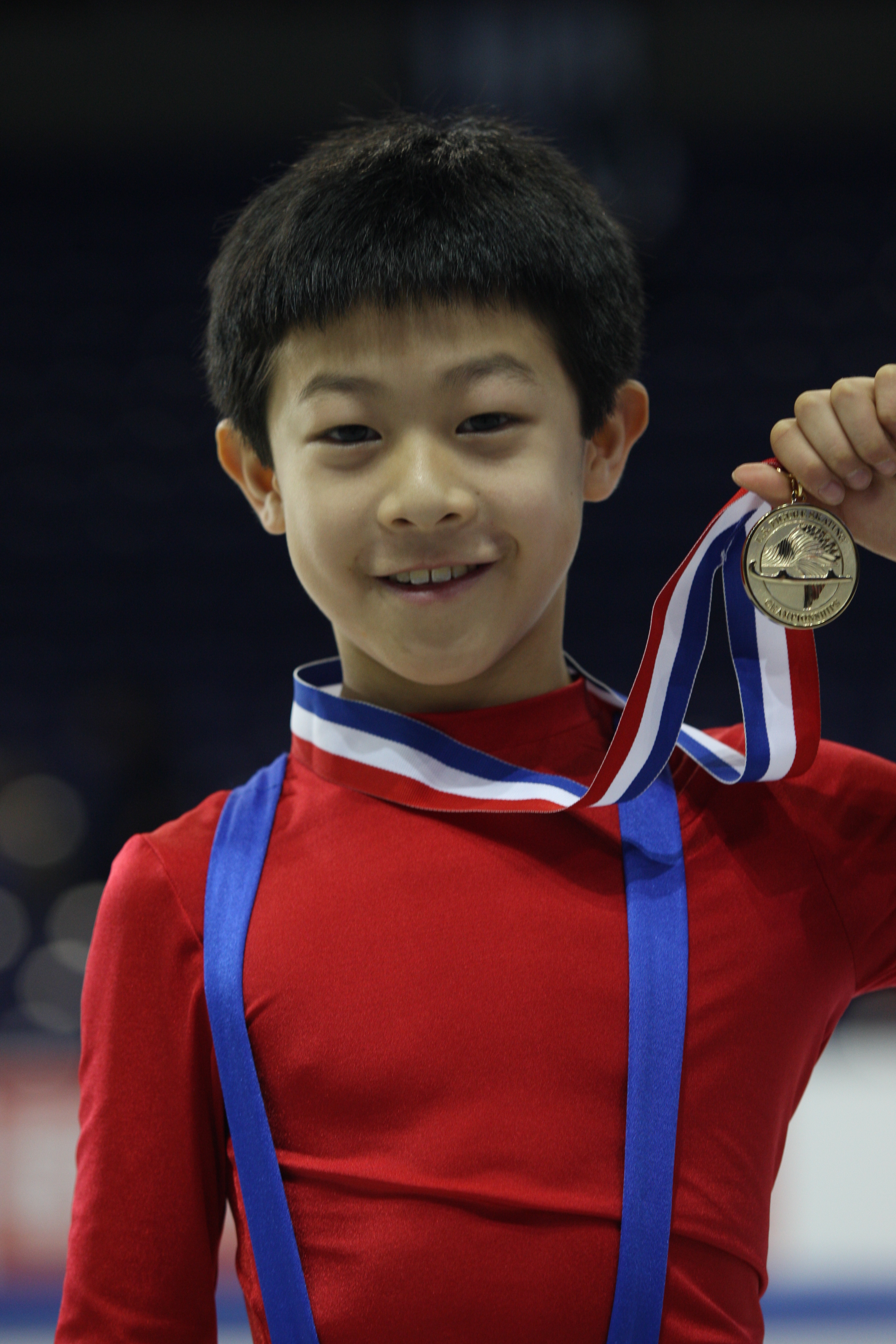 Nathan Chen as a young boy in red holds up a medal atop a podium. 